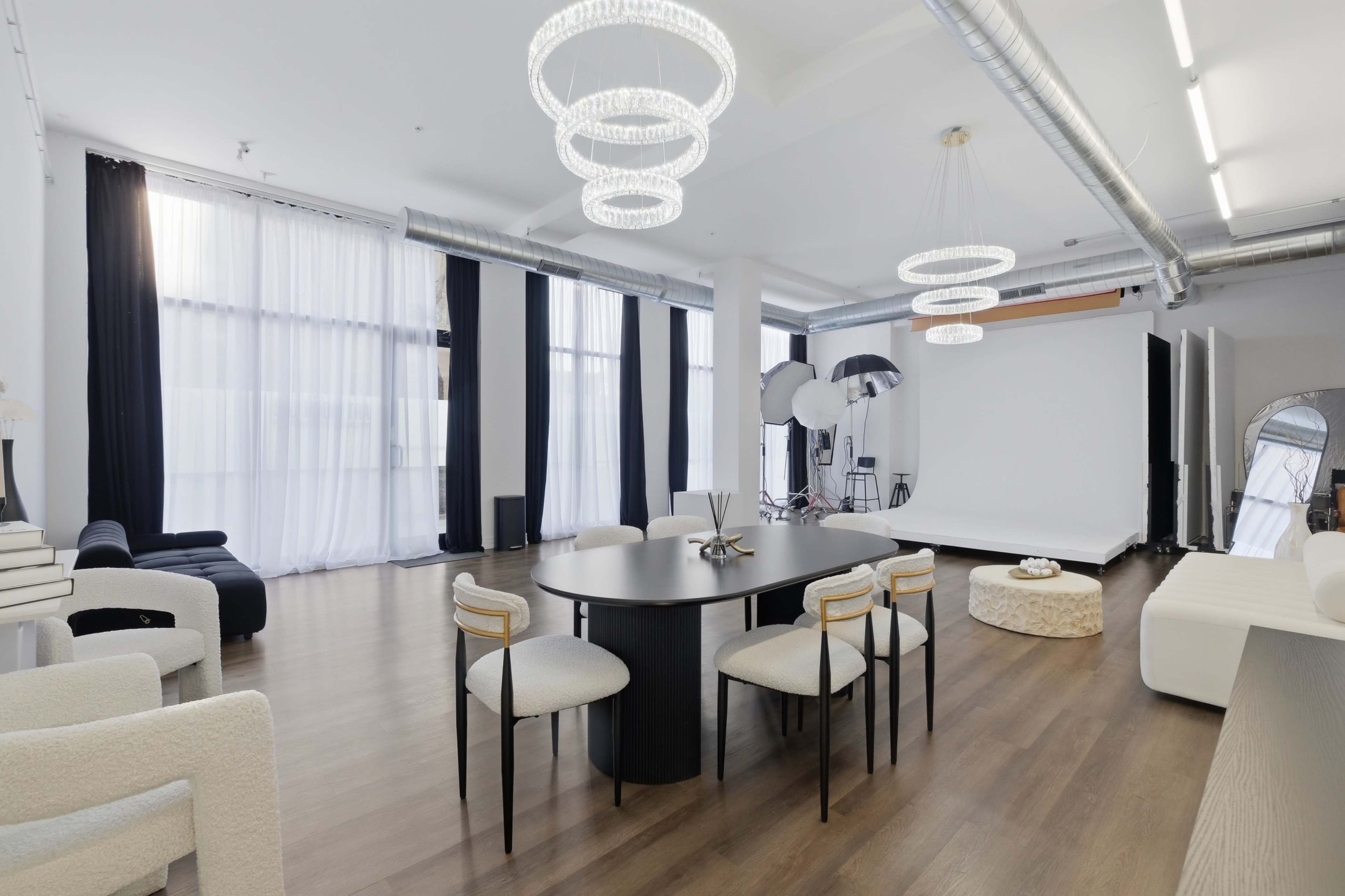 The image shows a spacious, modern interior with a dining area featuring a circular table and upholstered chairs, along with large windows and contemporary lighting.
