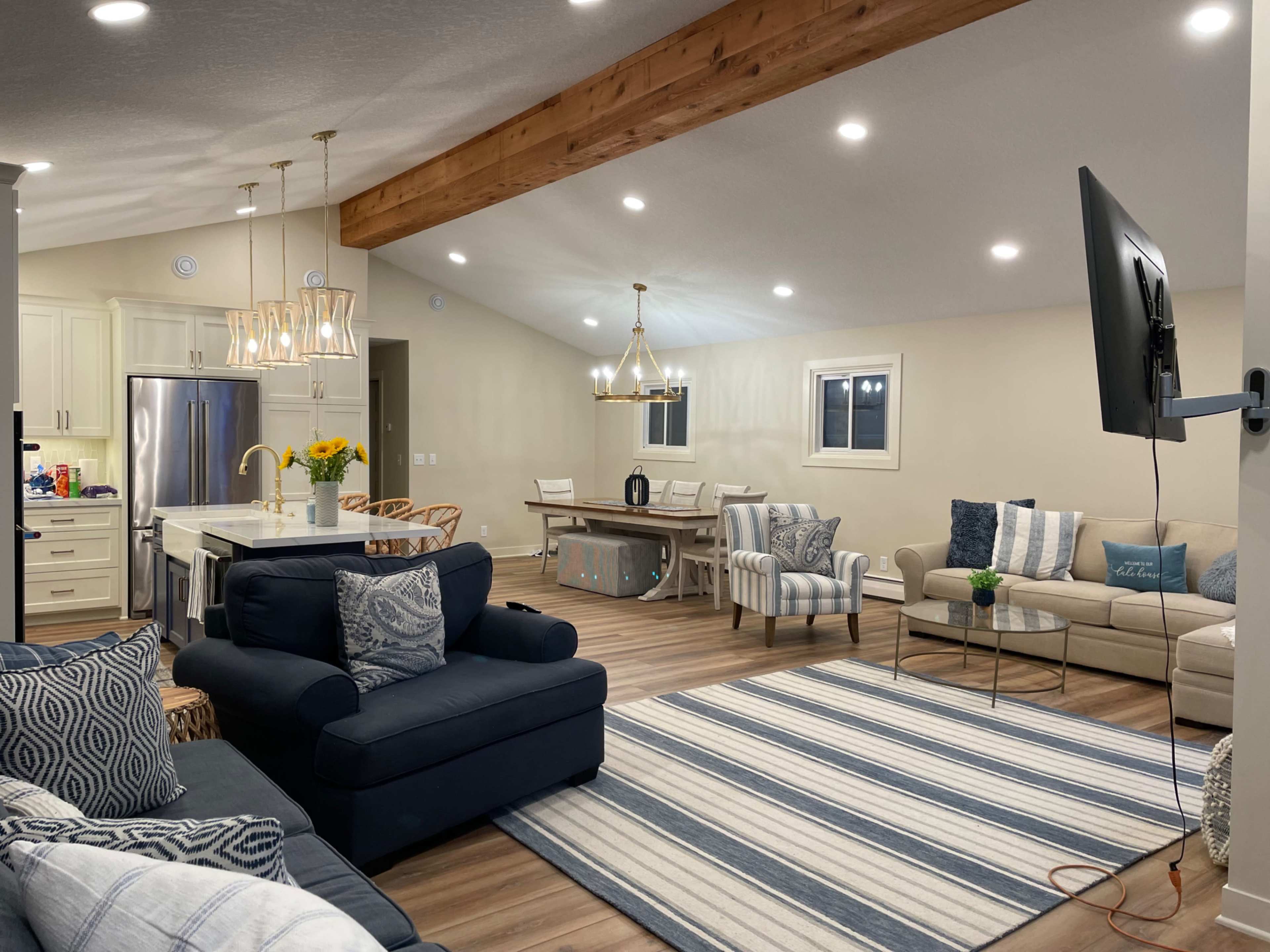 A modern living space featuring a combination of a kitchen and living area, with seating arrangements including sofas and a dining table under wooden beams and bright ceiling lights.