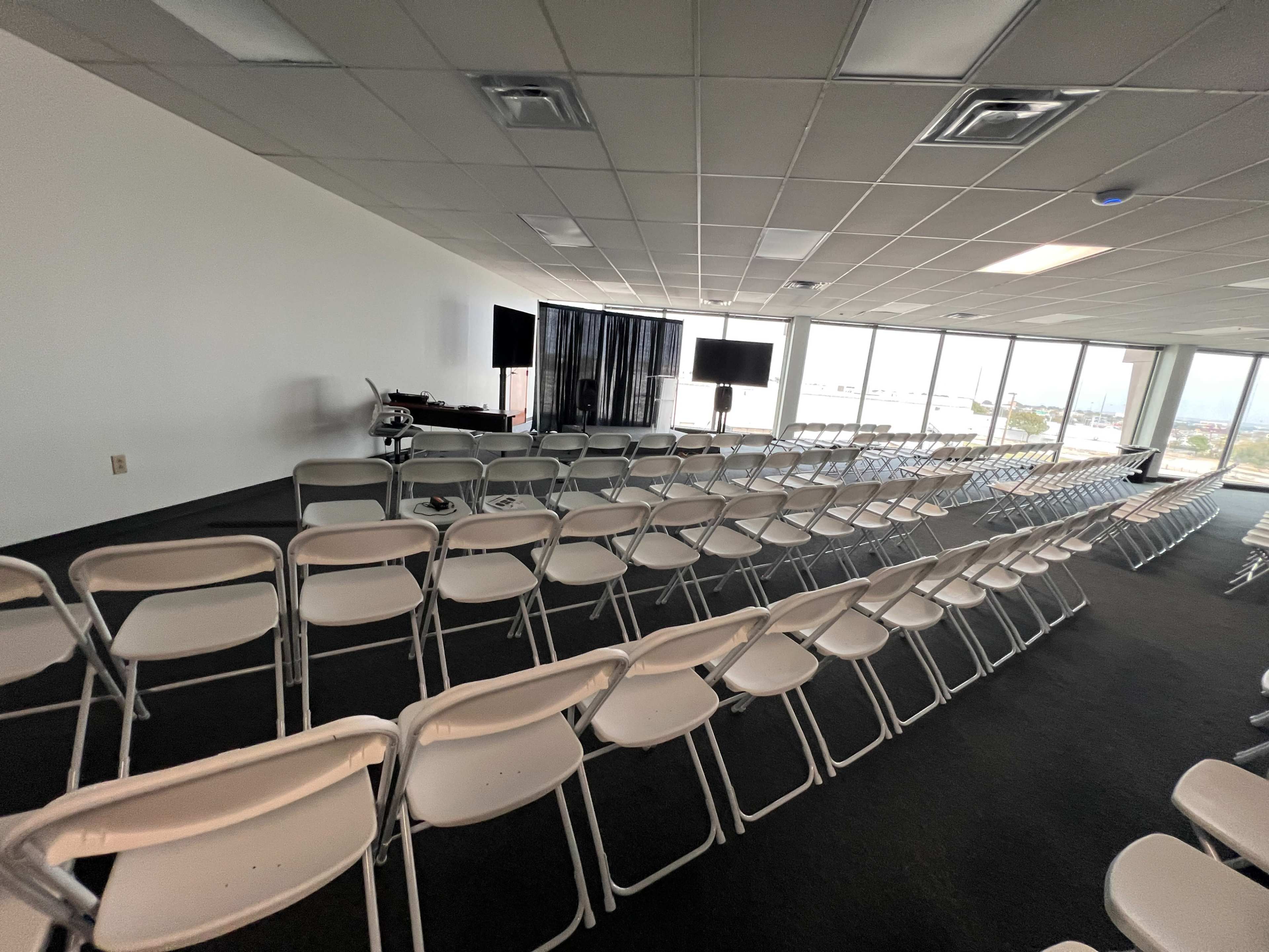 The image shows a spacious room arranged with rows of white folding chairs facing two screens, with large windows providing natural light.