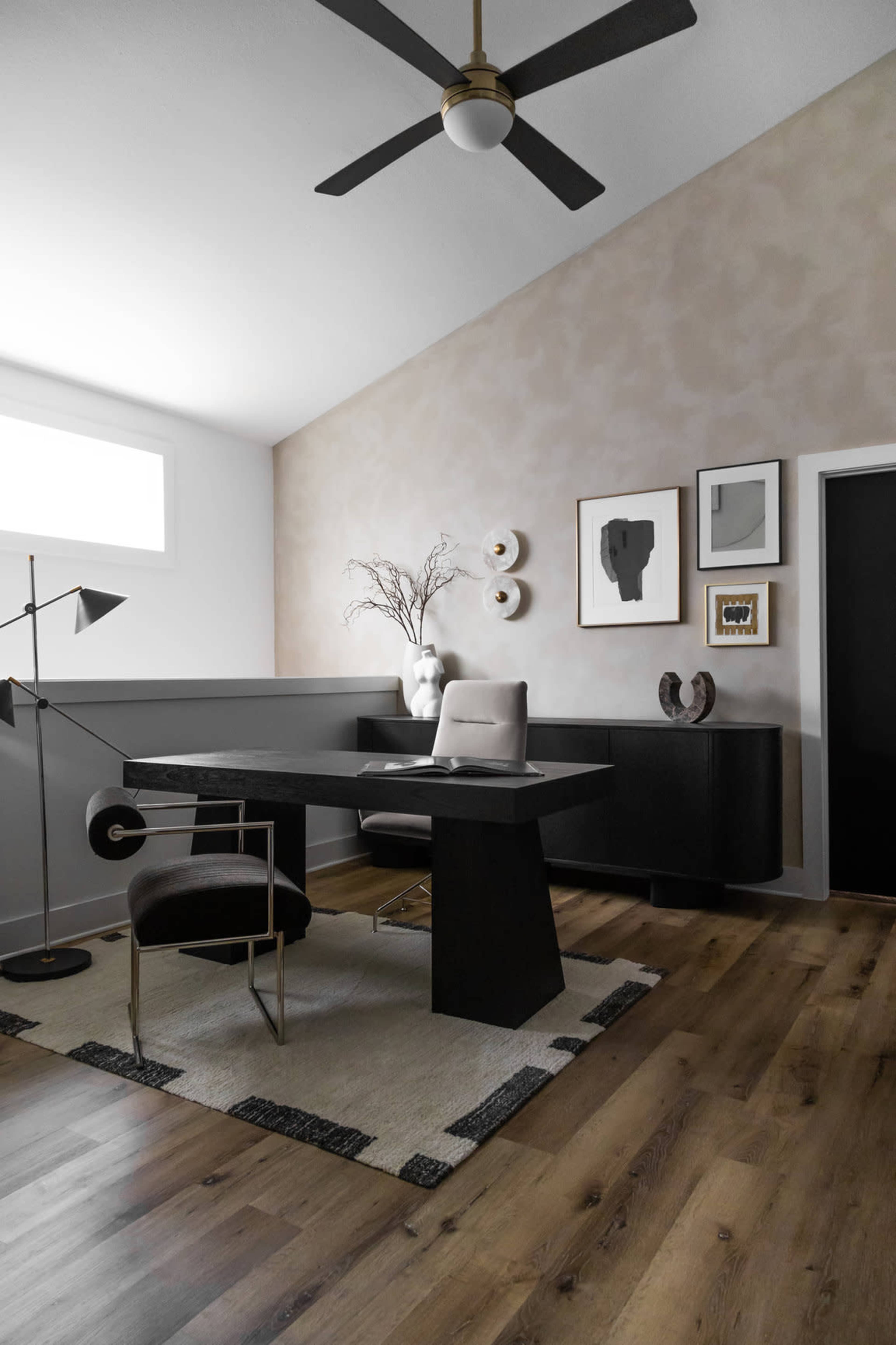 The image depicts a modern home office featuring a black desk, a white chair, and a stylish lamp, arranged on a light wood floor with a decorative rug.