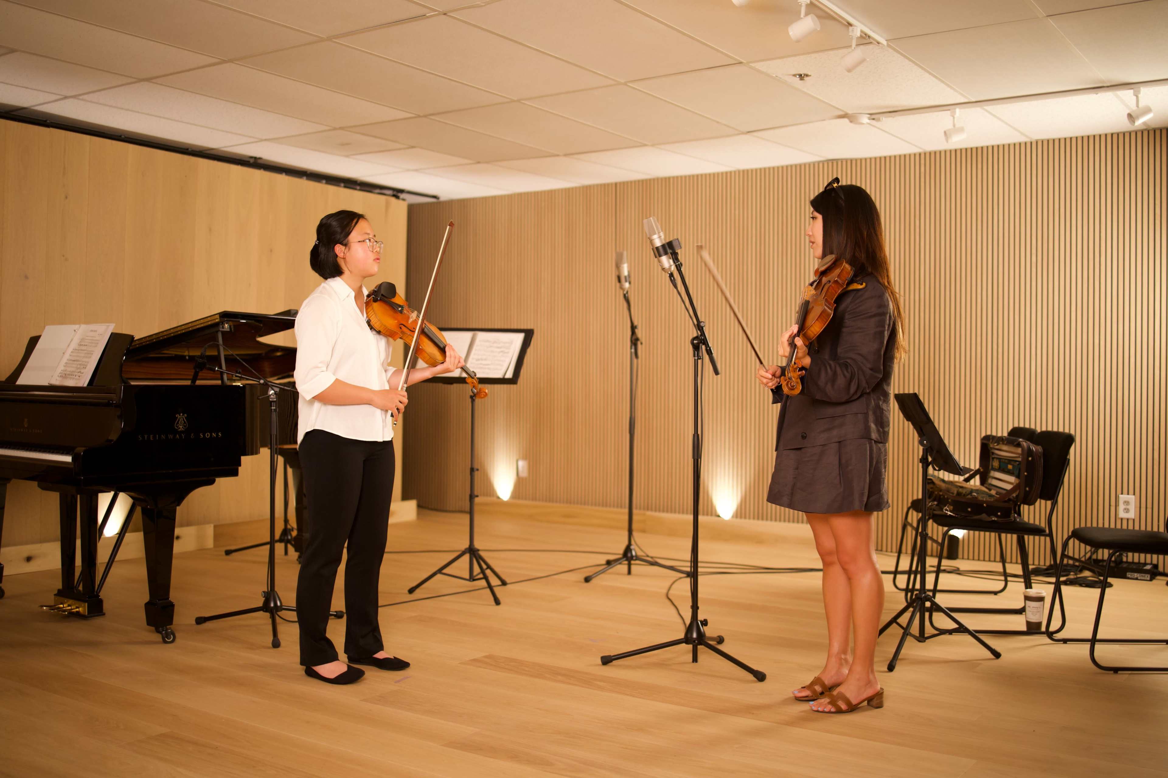 Two women stand in a music studio, each holding a violin, with sheet music and microphones set up in the background.