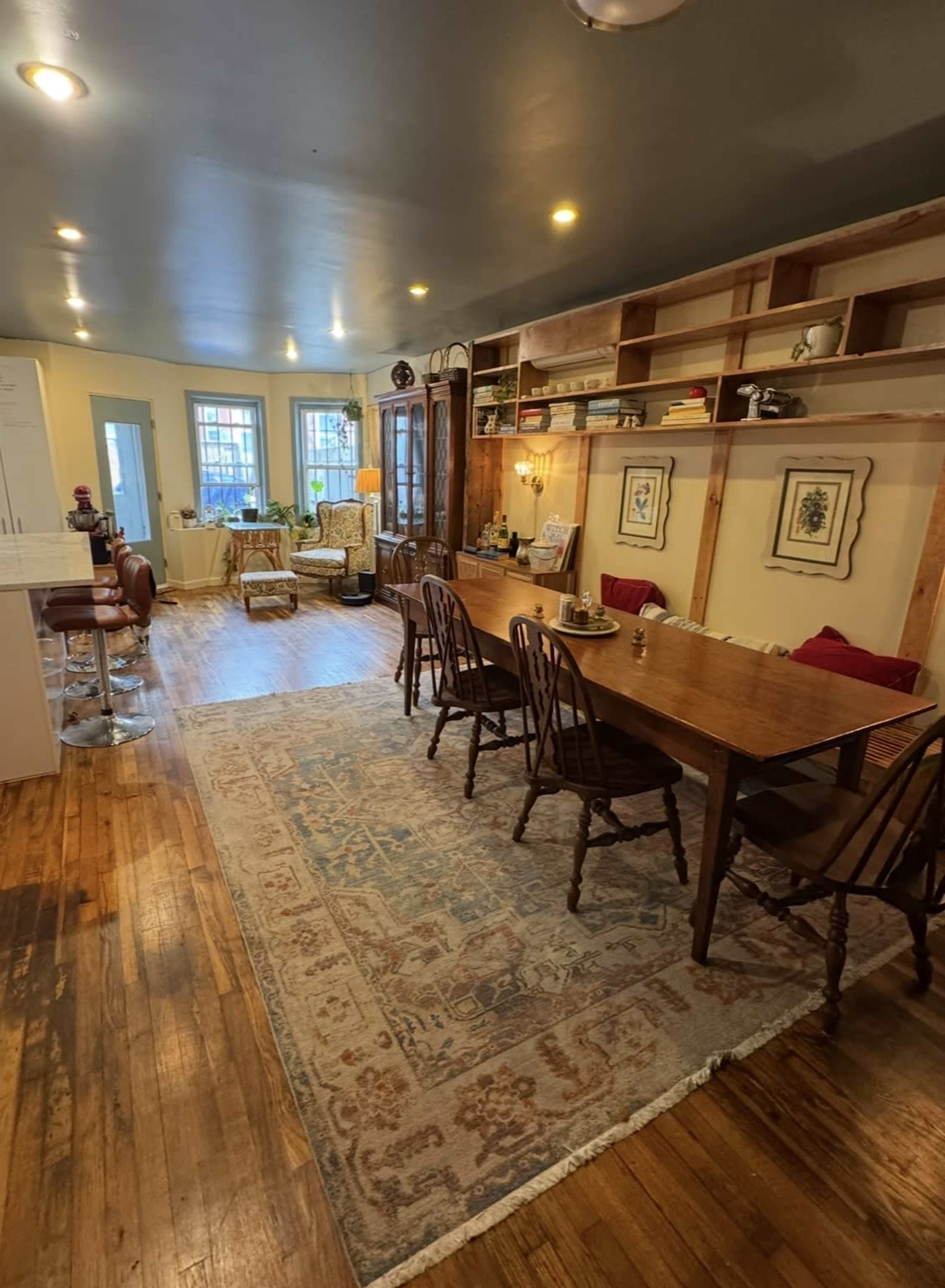 The image shows a spacious living area with a wooden dining table, chairs, and a cozy seating arrangement, illuminated by natural light from large windows.