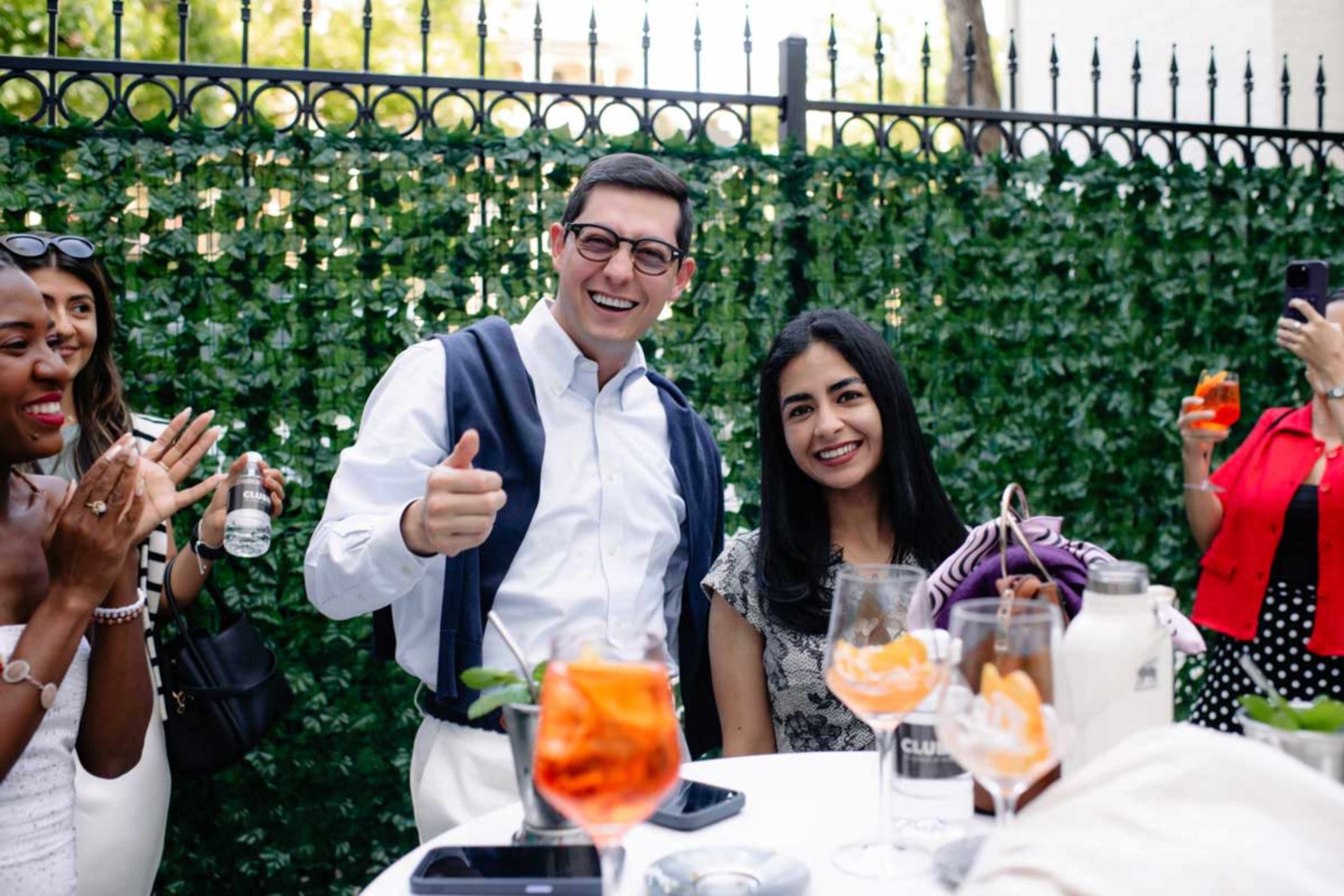 A group of people is gathered outdoors at a social event, with one man and one woman smiling and posing for the camera while others enjoy drinks in the background.