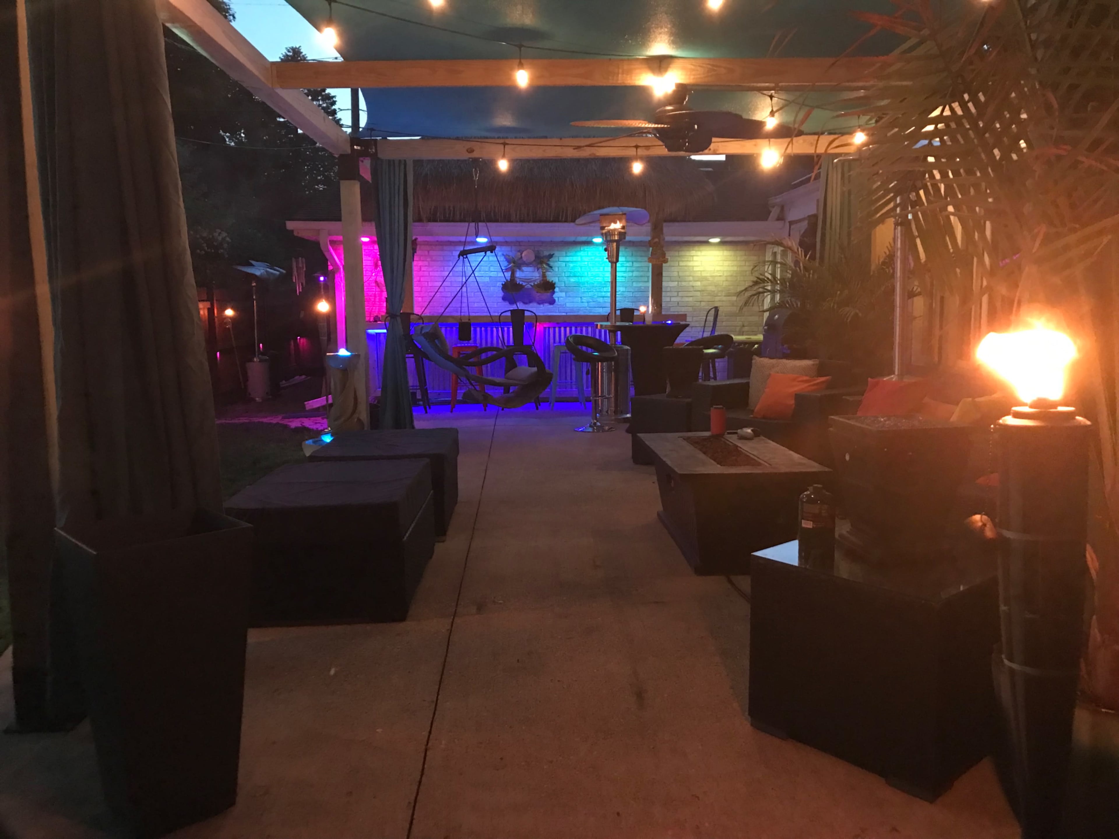 A backyard patio area illuminated by string lights, featuring seating arrangements with cushions, a table, and decorative elements like torches and colorful lighting.