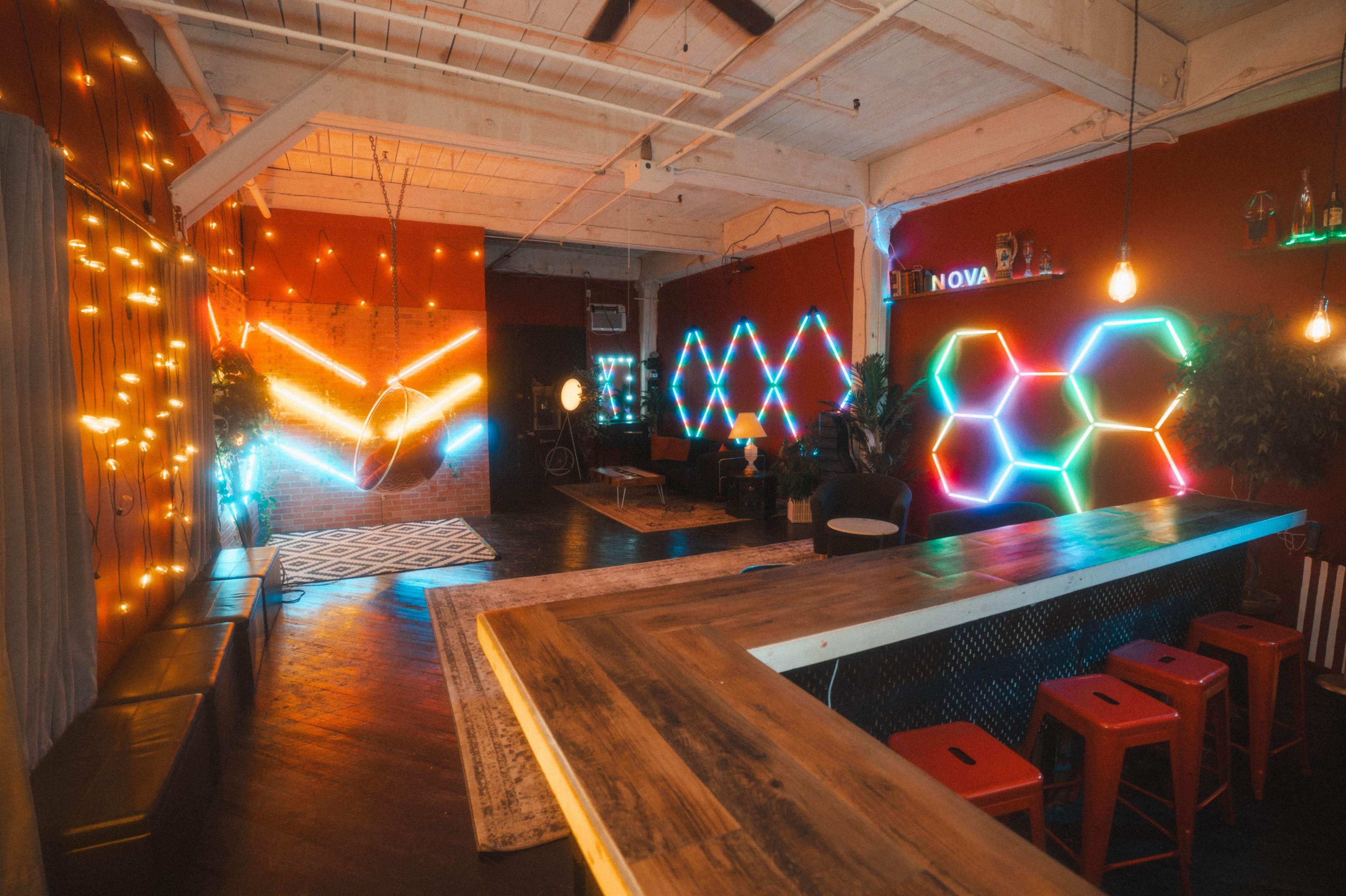 The image depicts a stylish interior space featuring colorful neon lights, a modern bar area, and a mix of seating options.
