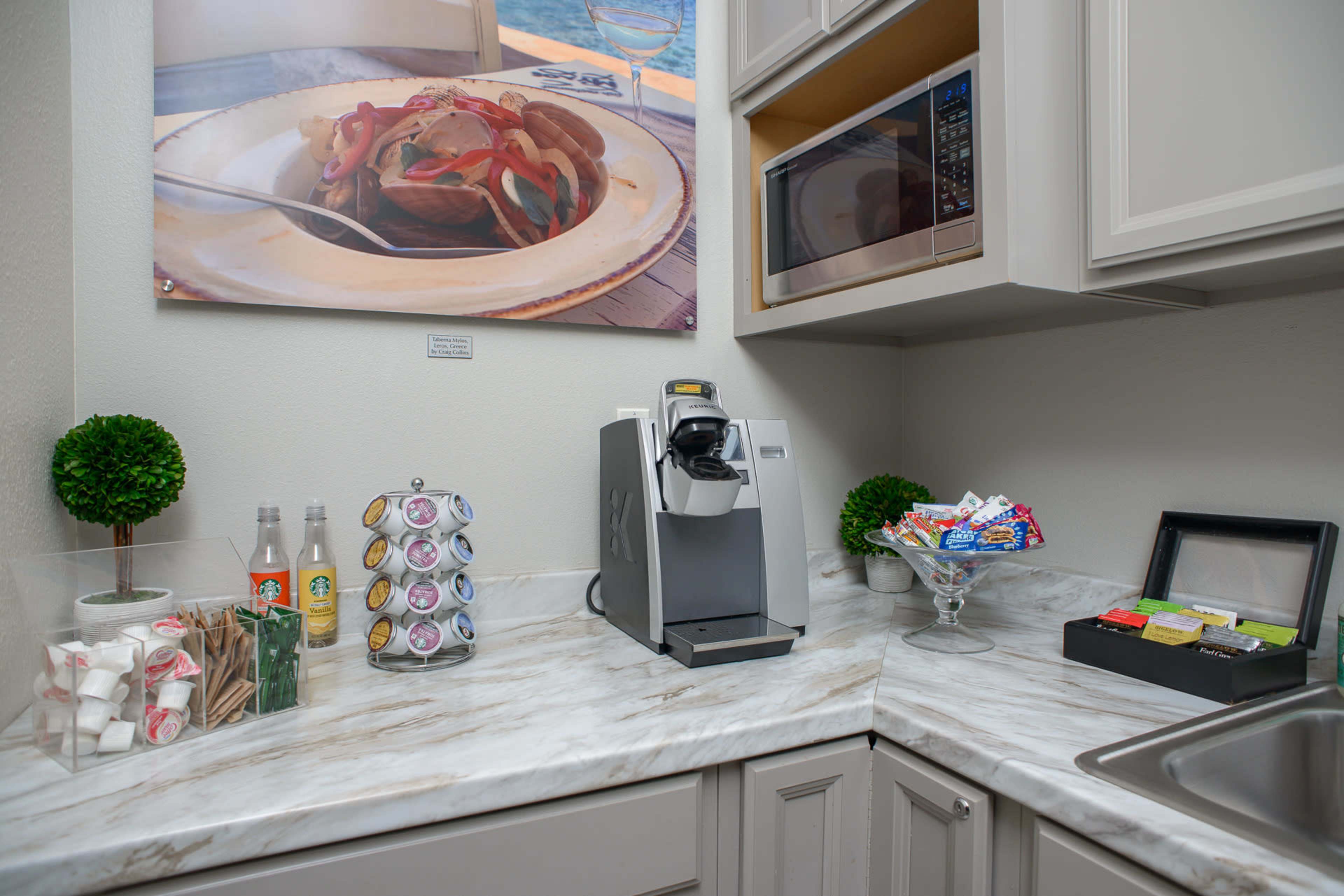 The image shows a small kitchen counter with a coffee maker, a microwave above, a bowl of snacks, and a display of drinks and utensils.