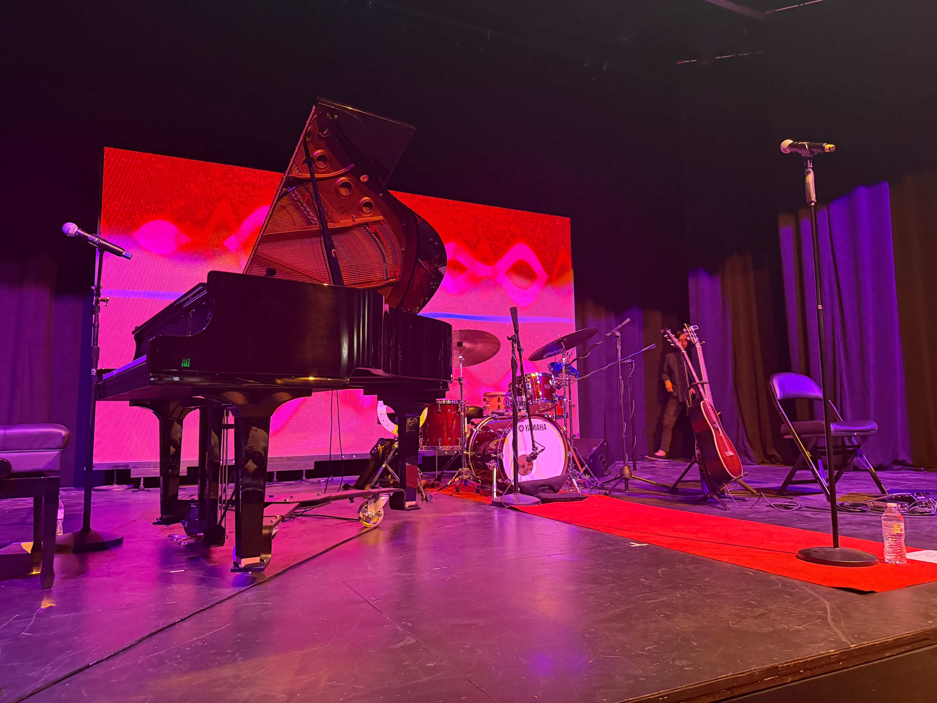 The stage is set with a grand piano, drums, a microphone stand, and a guitar, accompanied by a colorful background display.