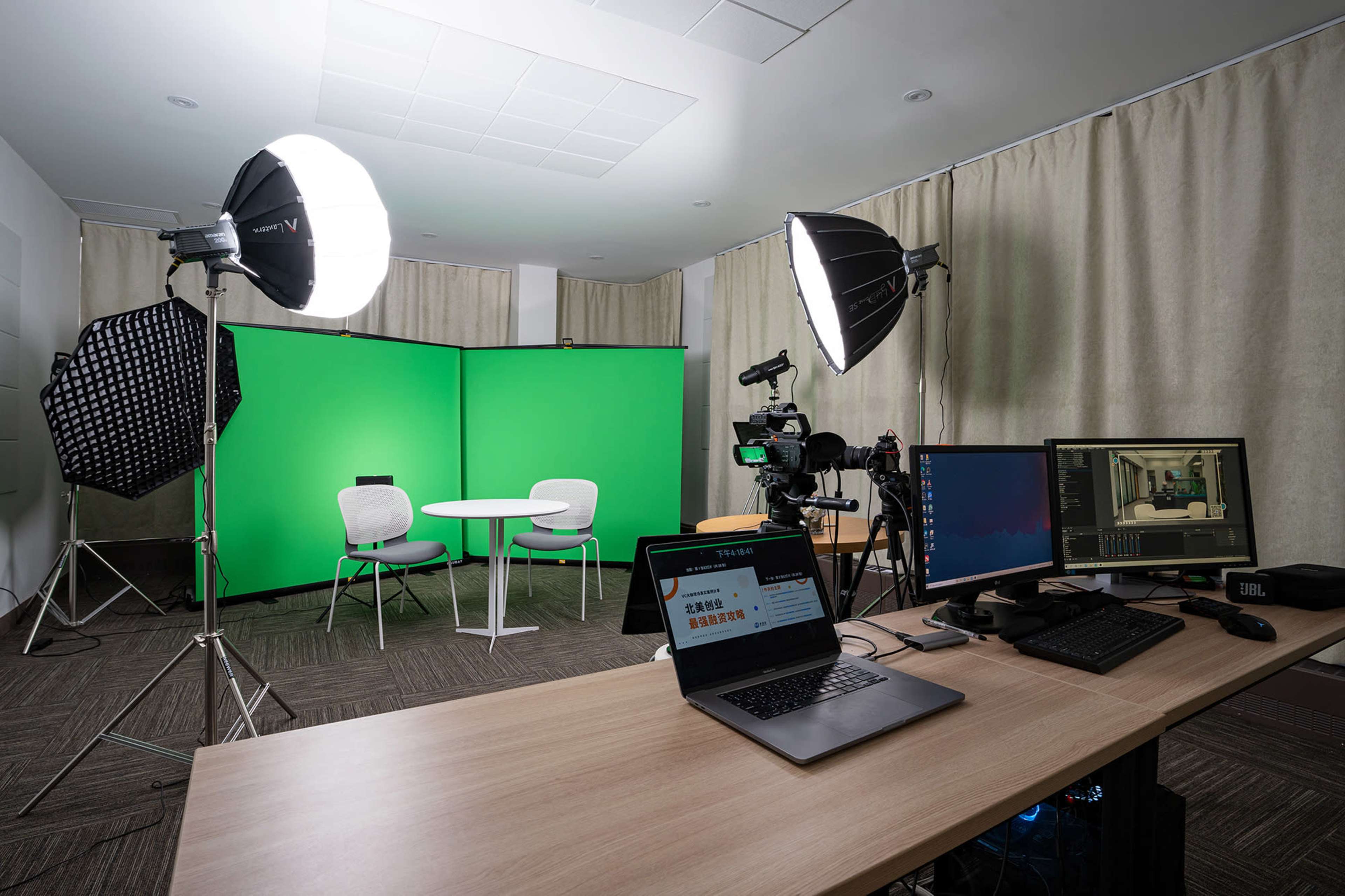 A filming studio features a green screen setup with cameras, lighting equipment, and a table with chairs.
