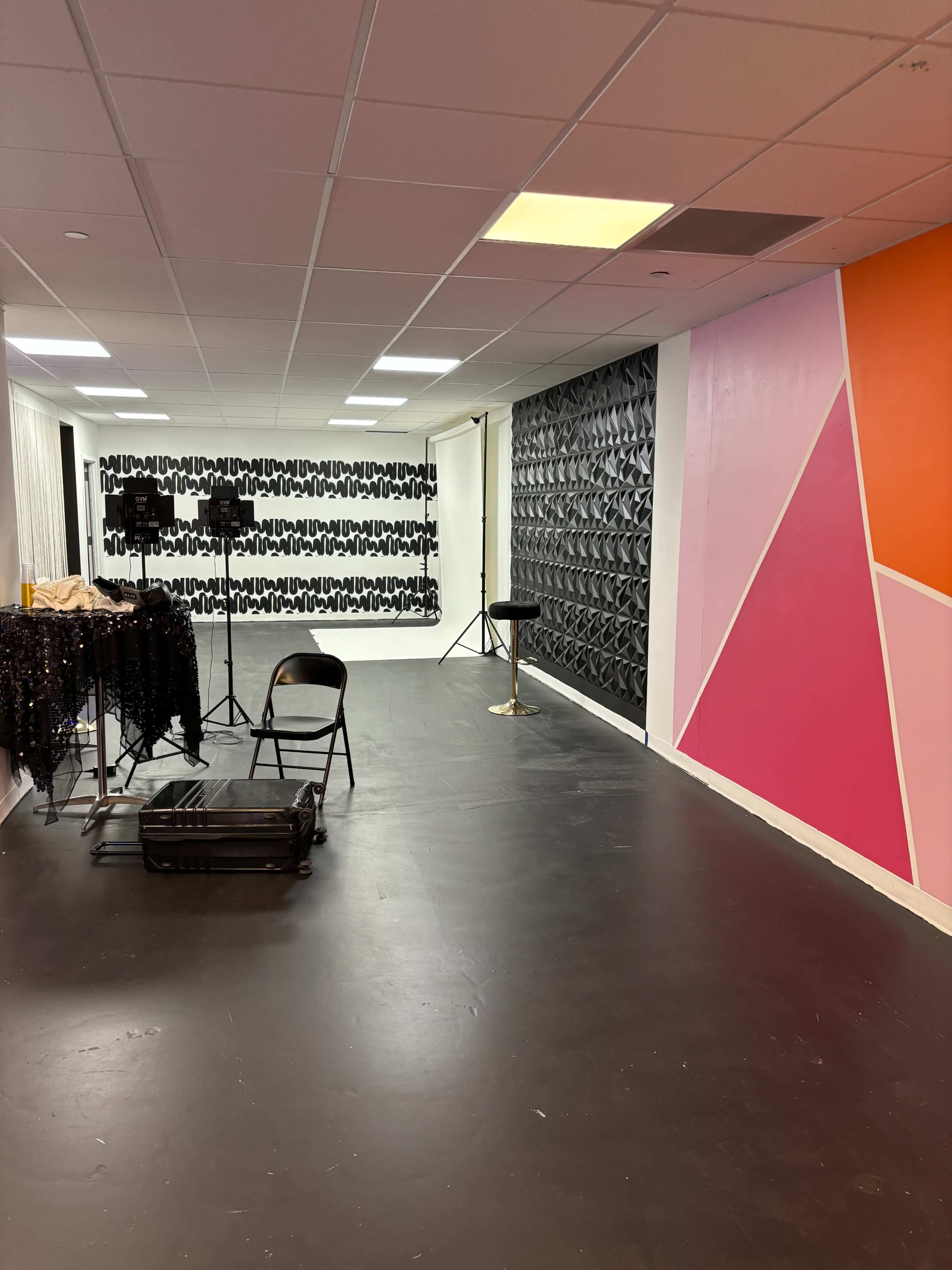 The image shows a spacious room with a black floor, featuring a patterned wall, a large solid-colored wall, and studio lighting equipment set up.