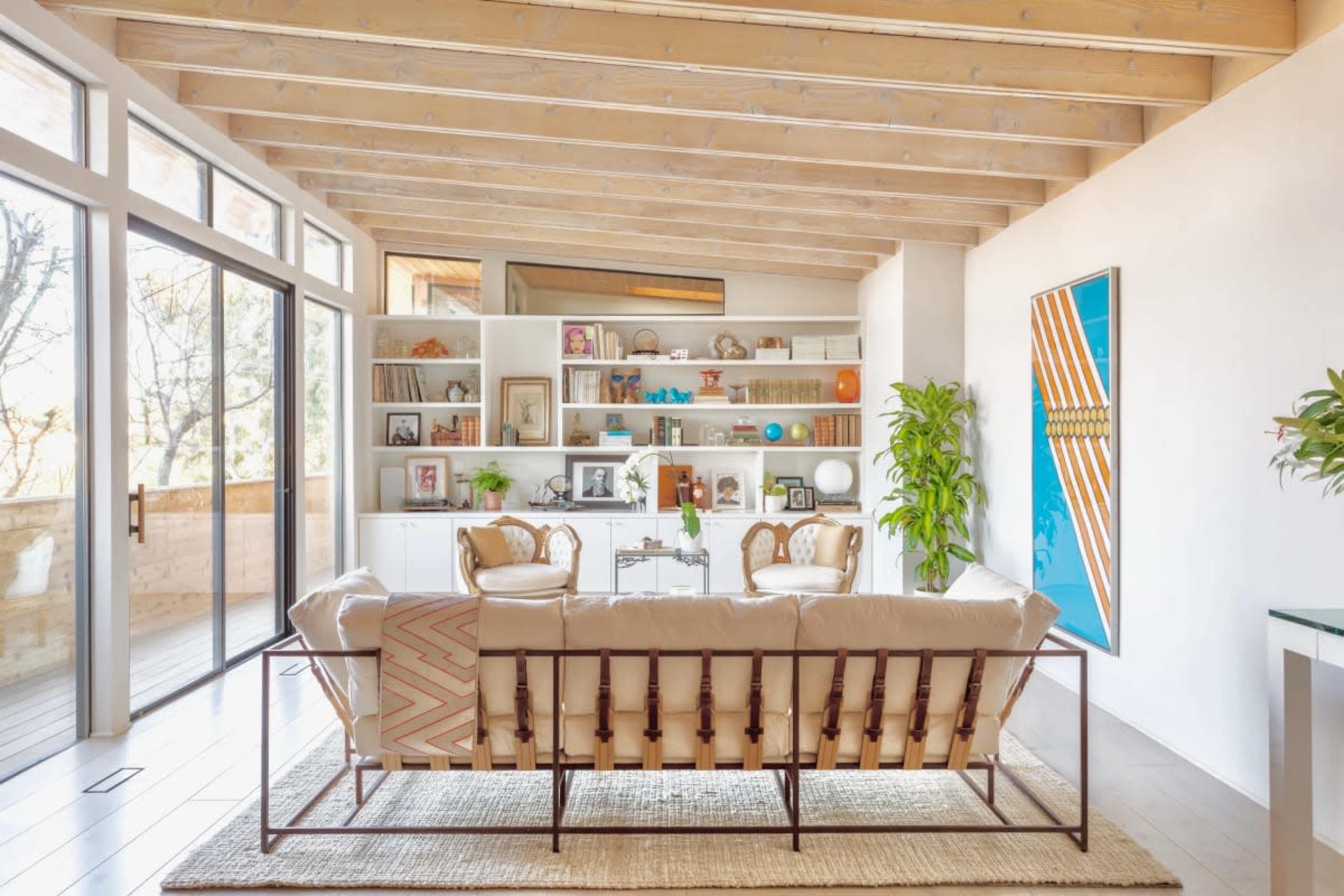 A modern living room features a beige sofa, two chairs, and a bookshelf filled with various decorative items, framed photos, and plants, with large windows allowing natural light.