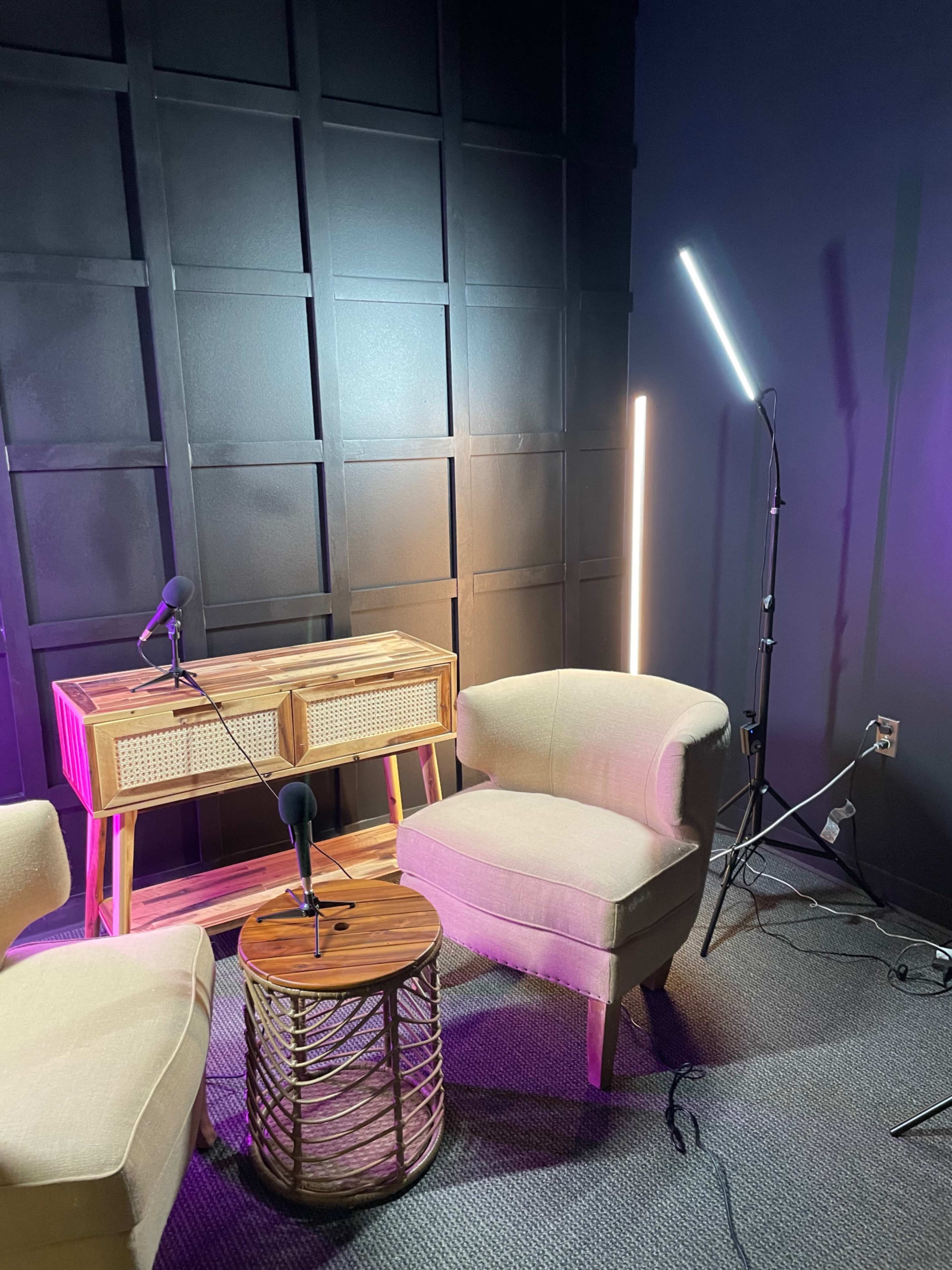 A small recording space featuring two beige chairs, a wooden table with a woven front, and studio lighting in a dark-colored room.