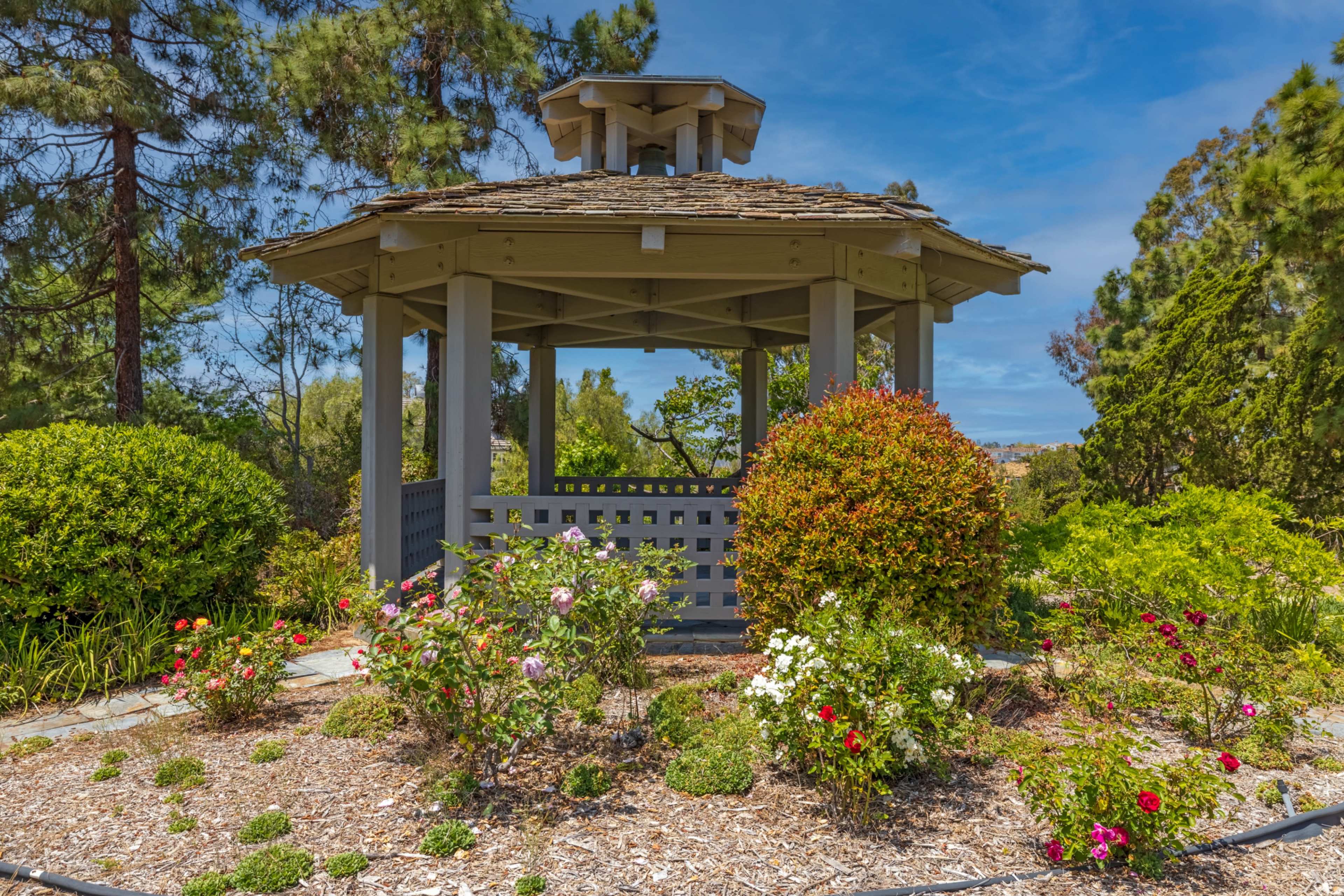 A wooden gazebo surrounded by flowering shrubs and decorative landscaping sits in a garden under a blue sky.