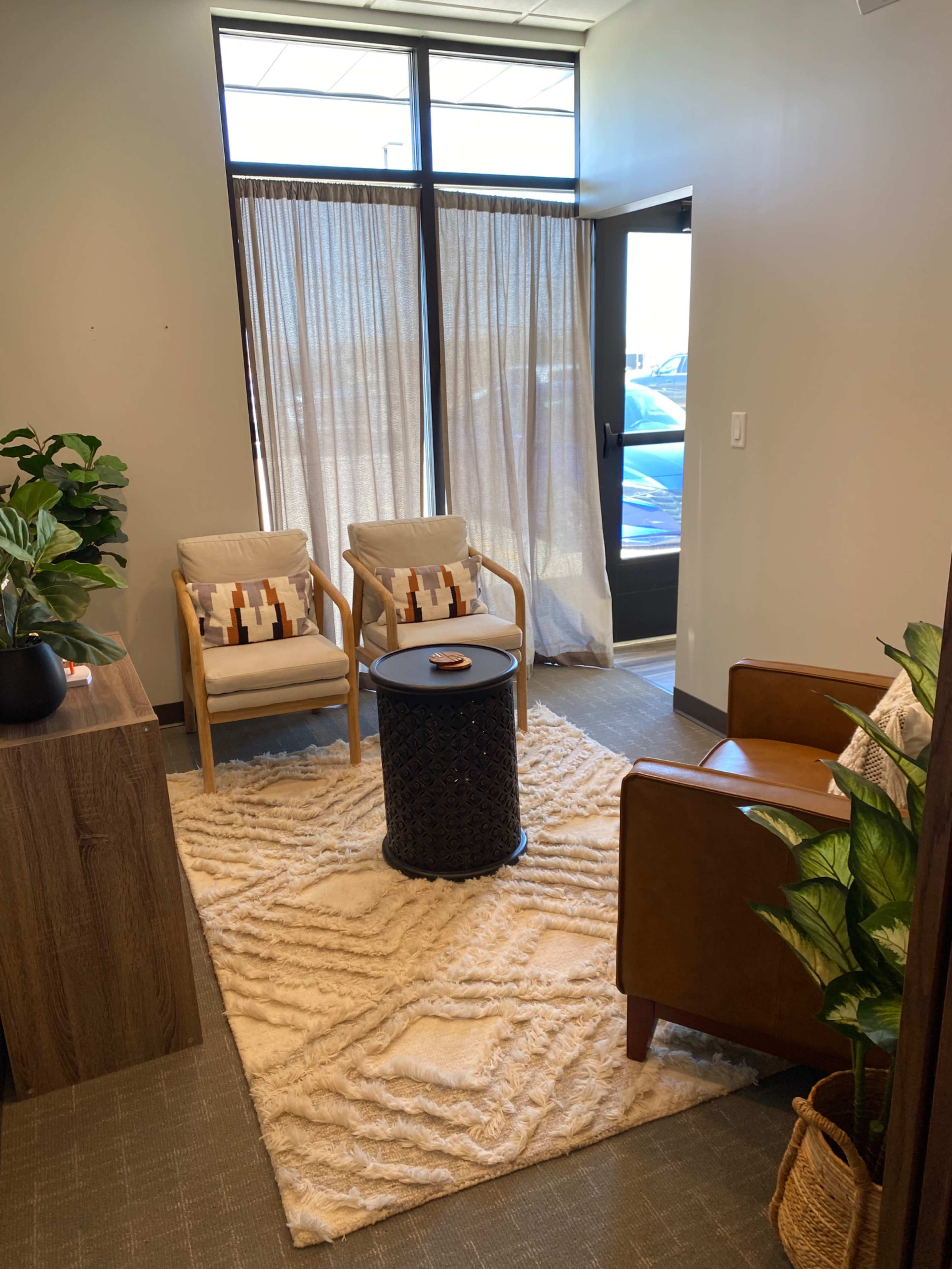 A small waiting area features two armchairs and a round table on a textured rug, with plants and light filtering through a sheer curtain.