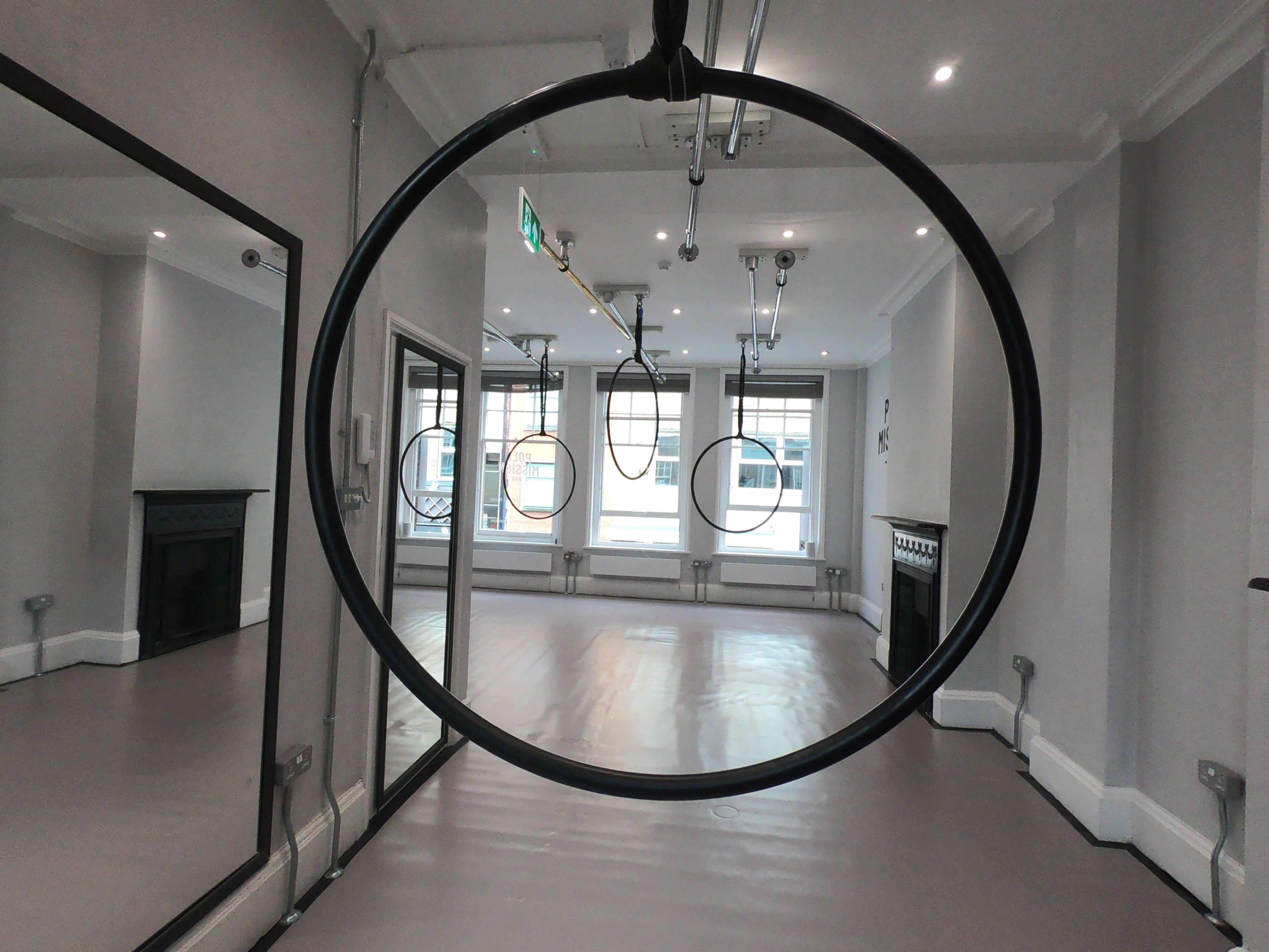 A spacious, well-lit room with large windows, a mirrored wall, and three circular fitness rings hanging from the ceiling.