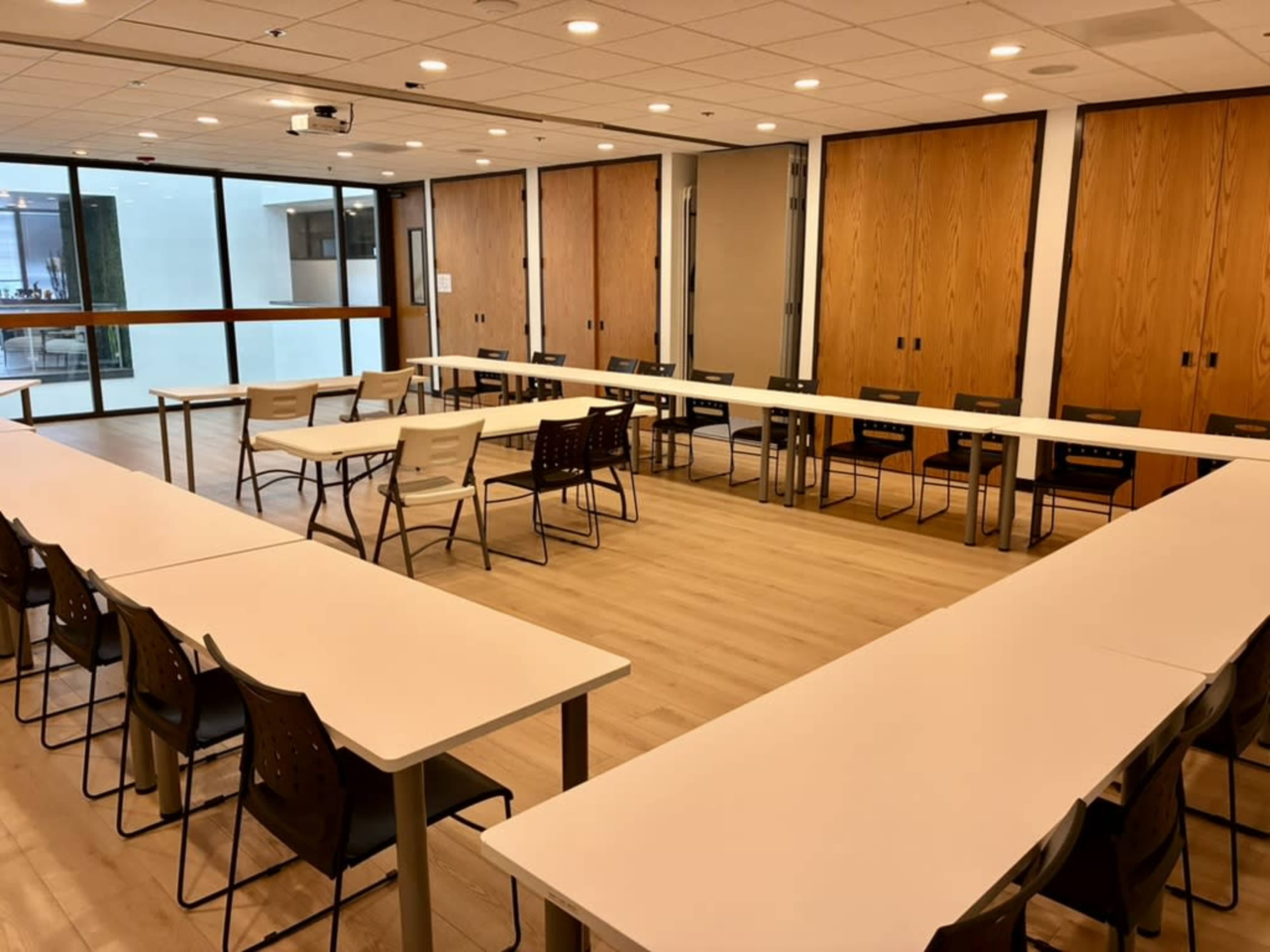 The image shows a meeting room arranged with several tables in a U-shape, surrounded by chairs, and featuring wooden doors and large windows.