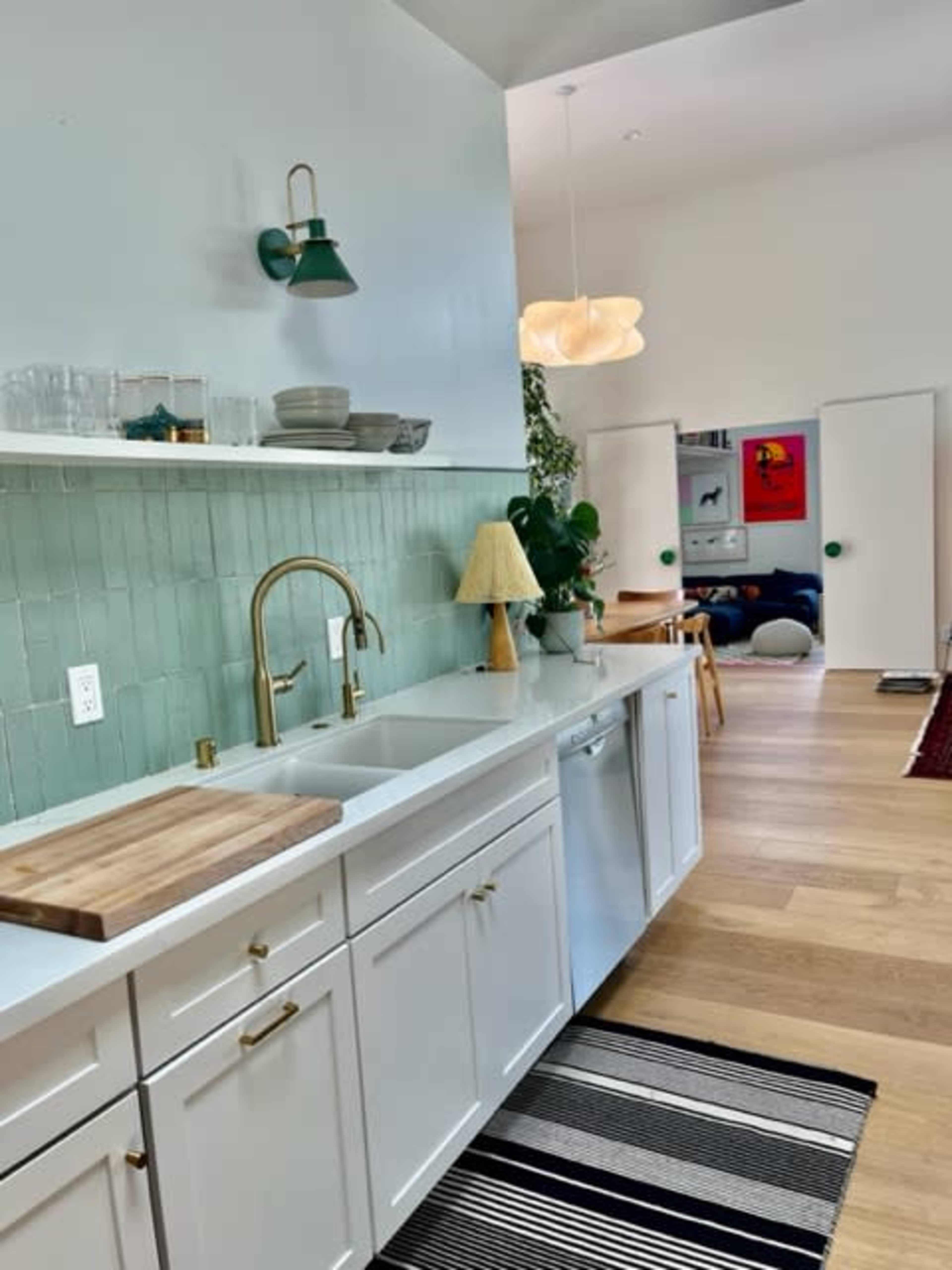 A modern kitchen features white cabinets, a green tiled backsplash, a wooden cutting board, and a pendant light, with a dishwasher and a plant on the counter.