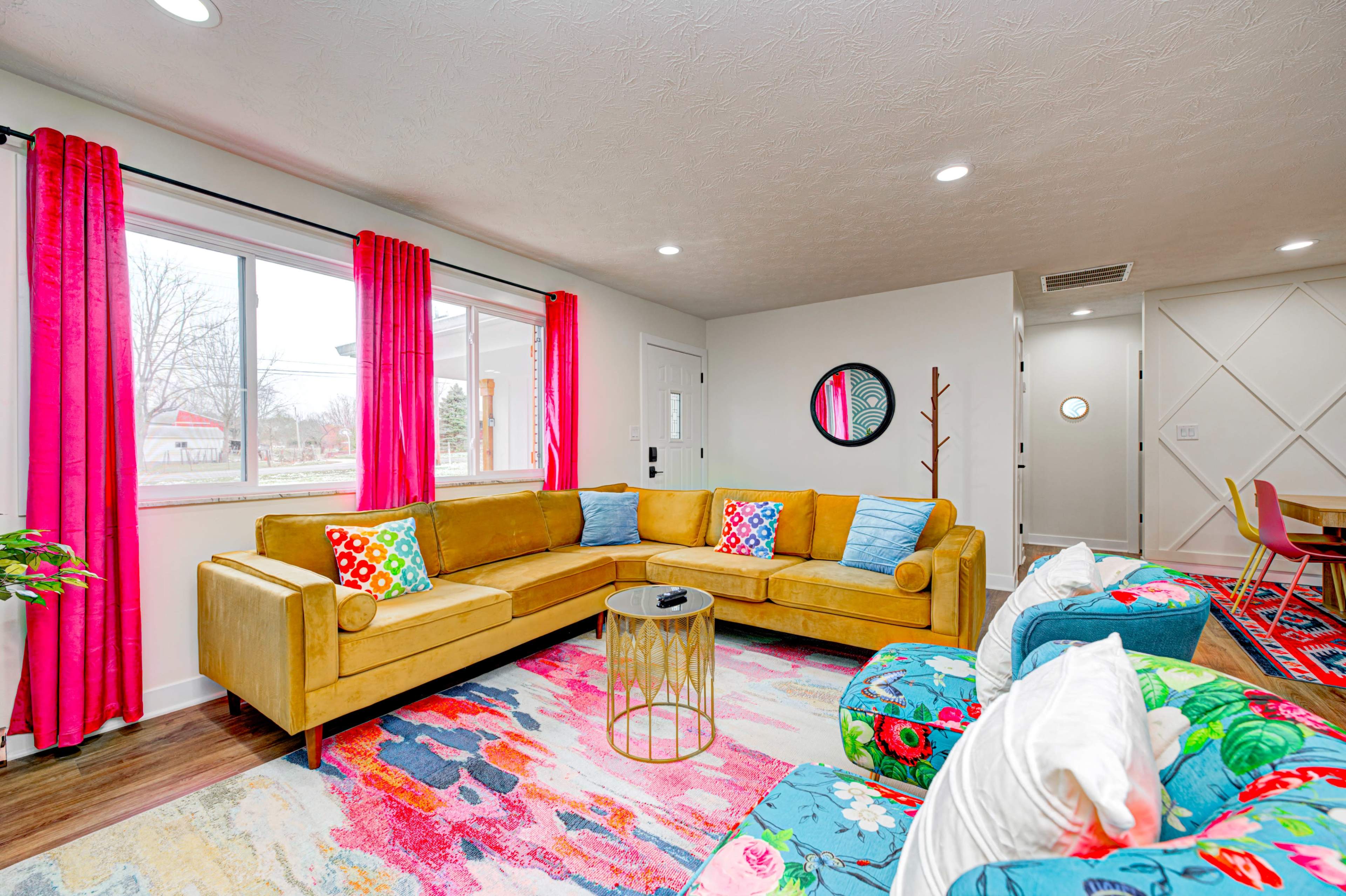 The image shows a vibrant living room featuring a mustard-colored sectional sofa, colorful armchairs, bright curtains, and a patterned rug, along with a dining area visible in the background.