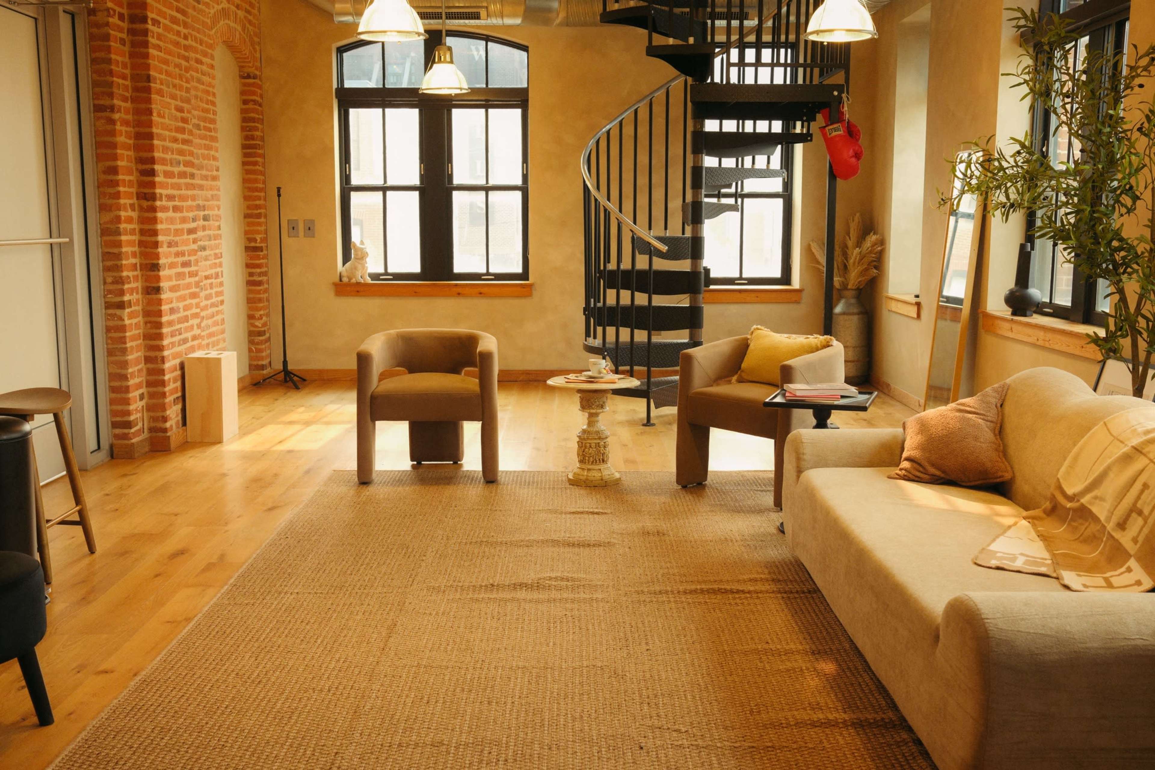 The image shows a modern living space with a spiral staircase, two armchairs, a sofa, and large windows, featuring wooden flooring and exposed brick walls.