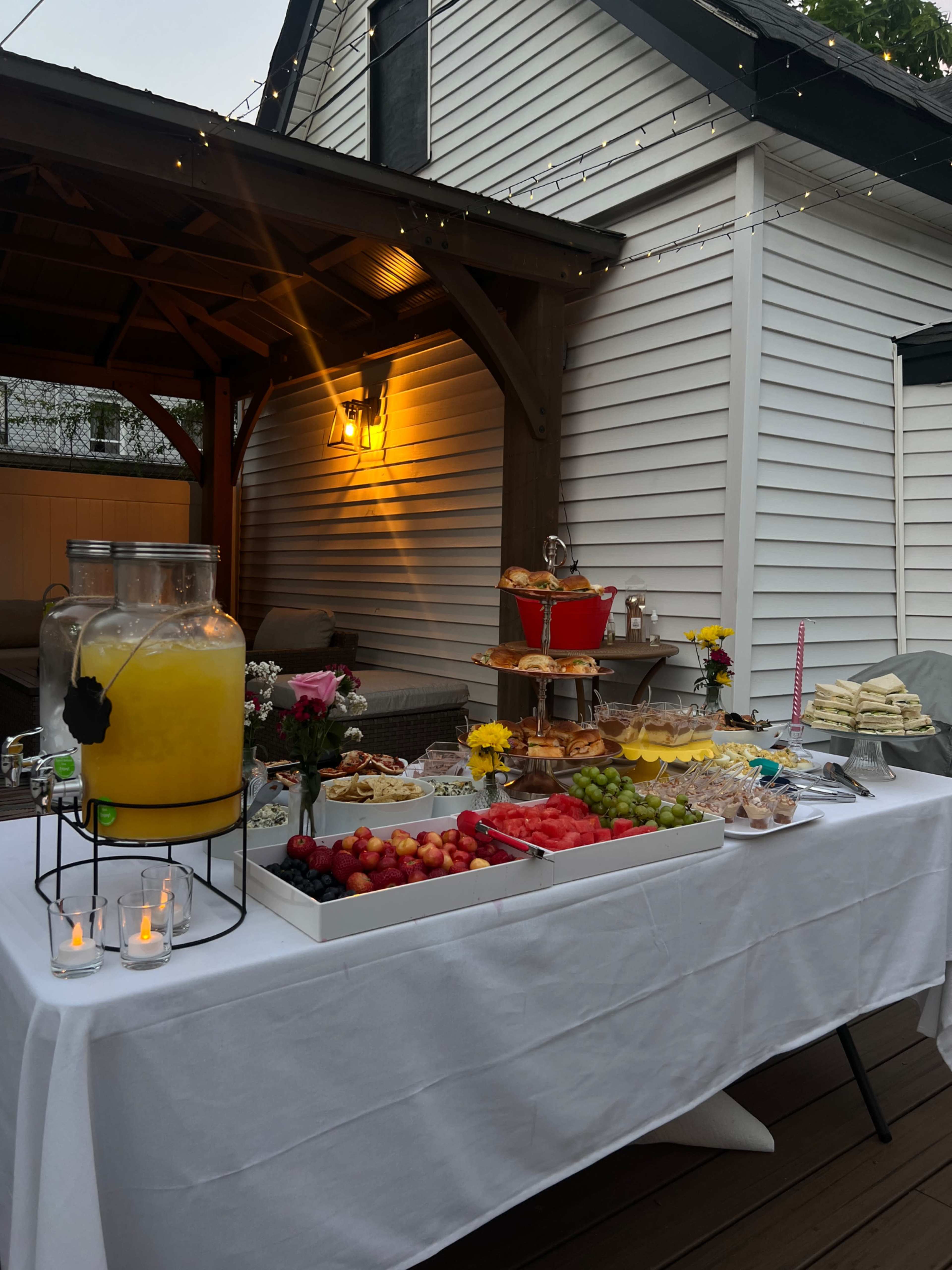A table is set outdoors under a gazebo, displaying a variety of food and beverages, including a large glass pitcher of yellow drink, fruits, pastries, and decorative flowers.