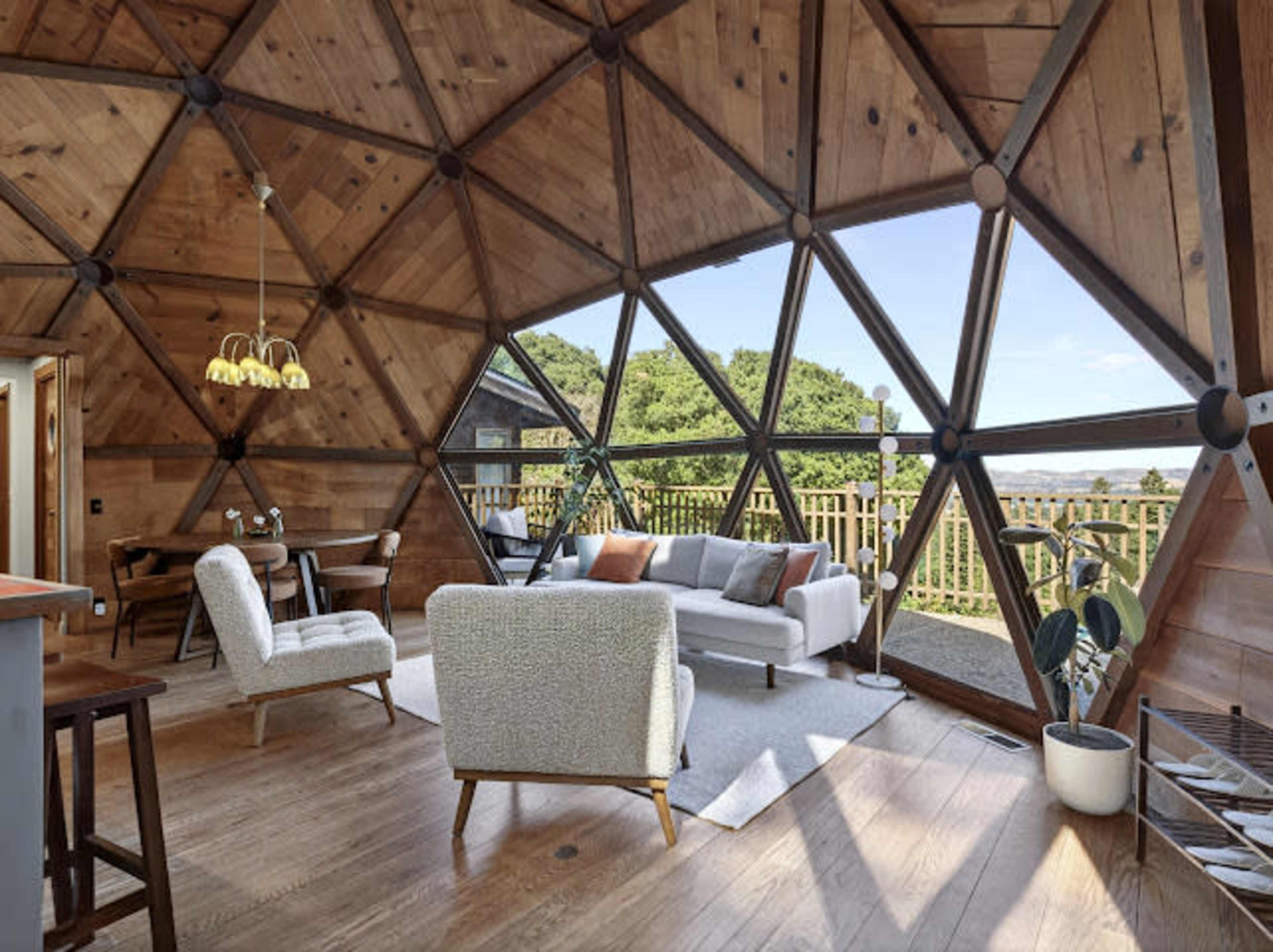 The image shows a geodesic dome interior featuring wooden walls, a comfortable seating area with a sofa and chairs, and large windows that provide a view of greenery outside.