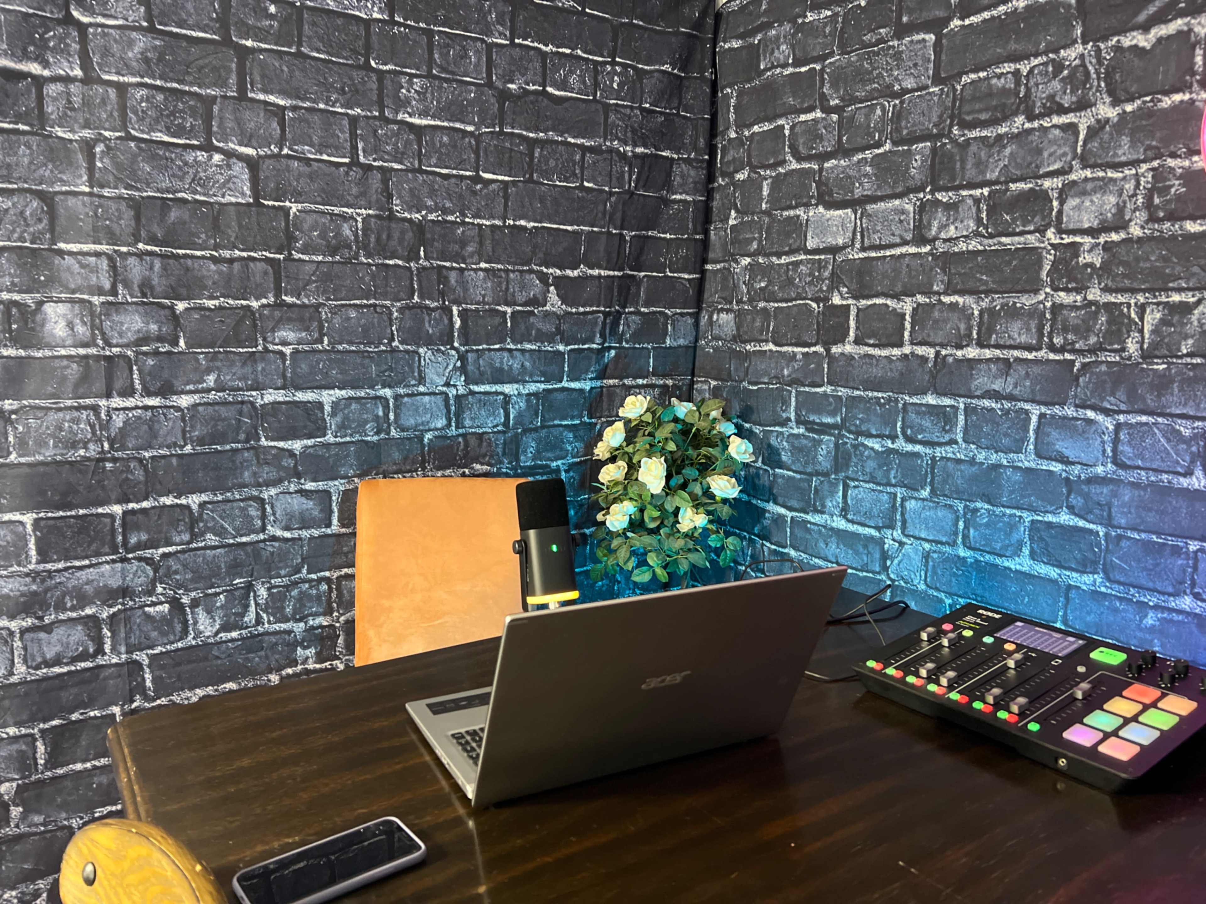 A wooden table with a laptop, microphone, and music mixer is set against a backdrop of black brick walls and a small plant.