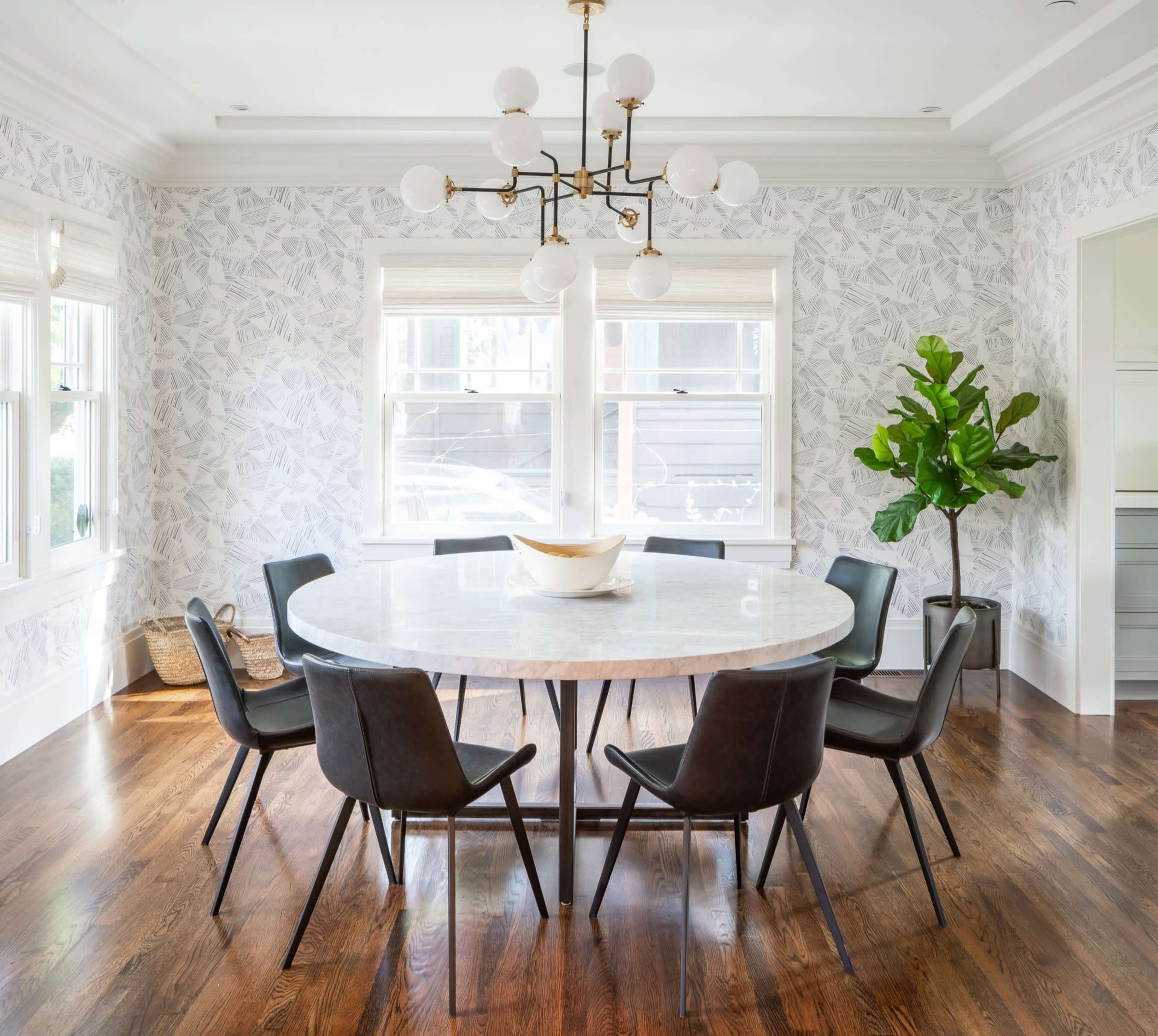 A round dining table surrounded by black chairs is set in a well-lit room with patterned wallpaper and a large plant in the corner.