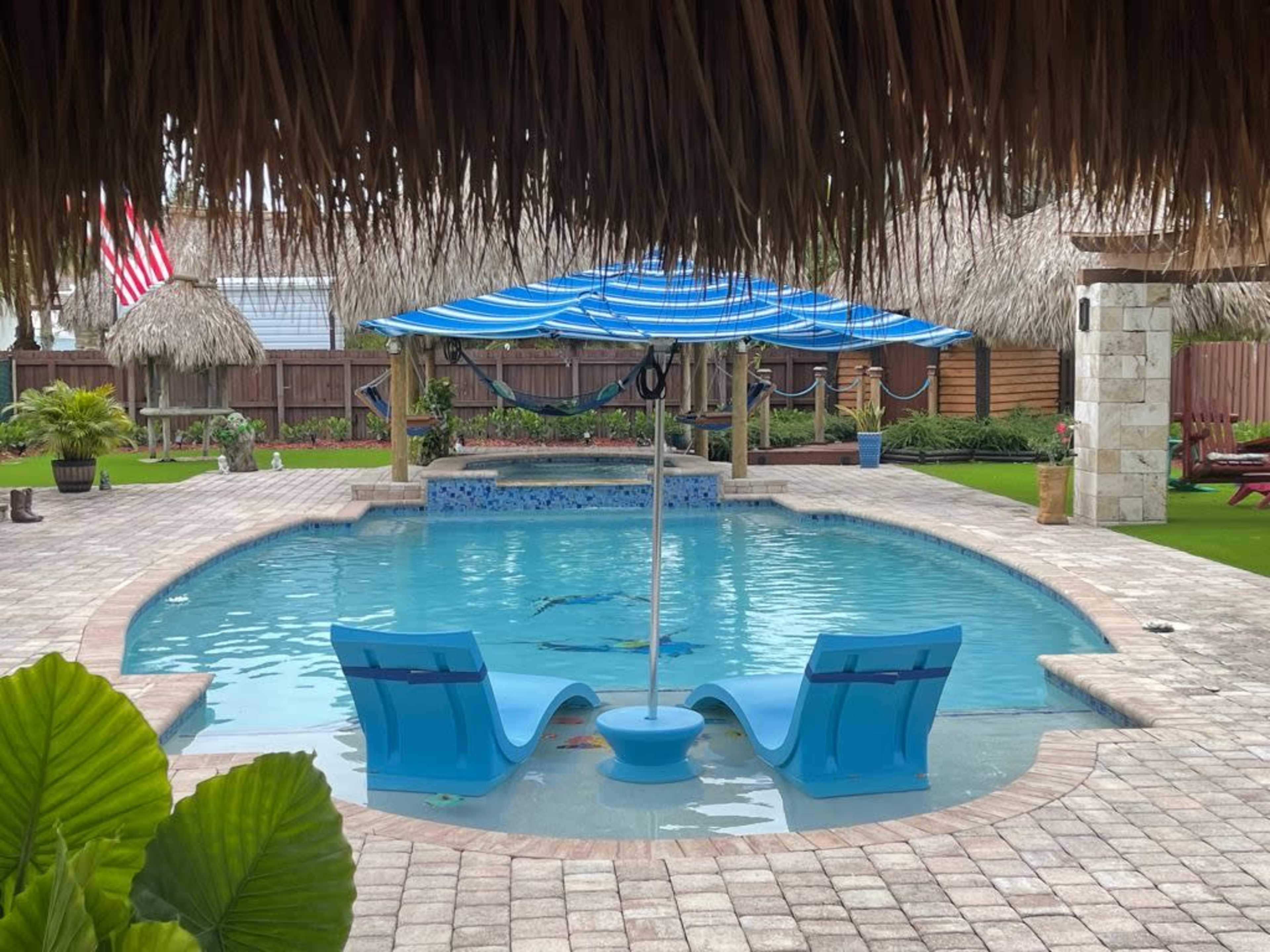 A swimming pool with a shaded seating area features two blue lounge chairs and an umbrella, surrounded by tropical landscaping and a thatched roof structure.