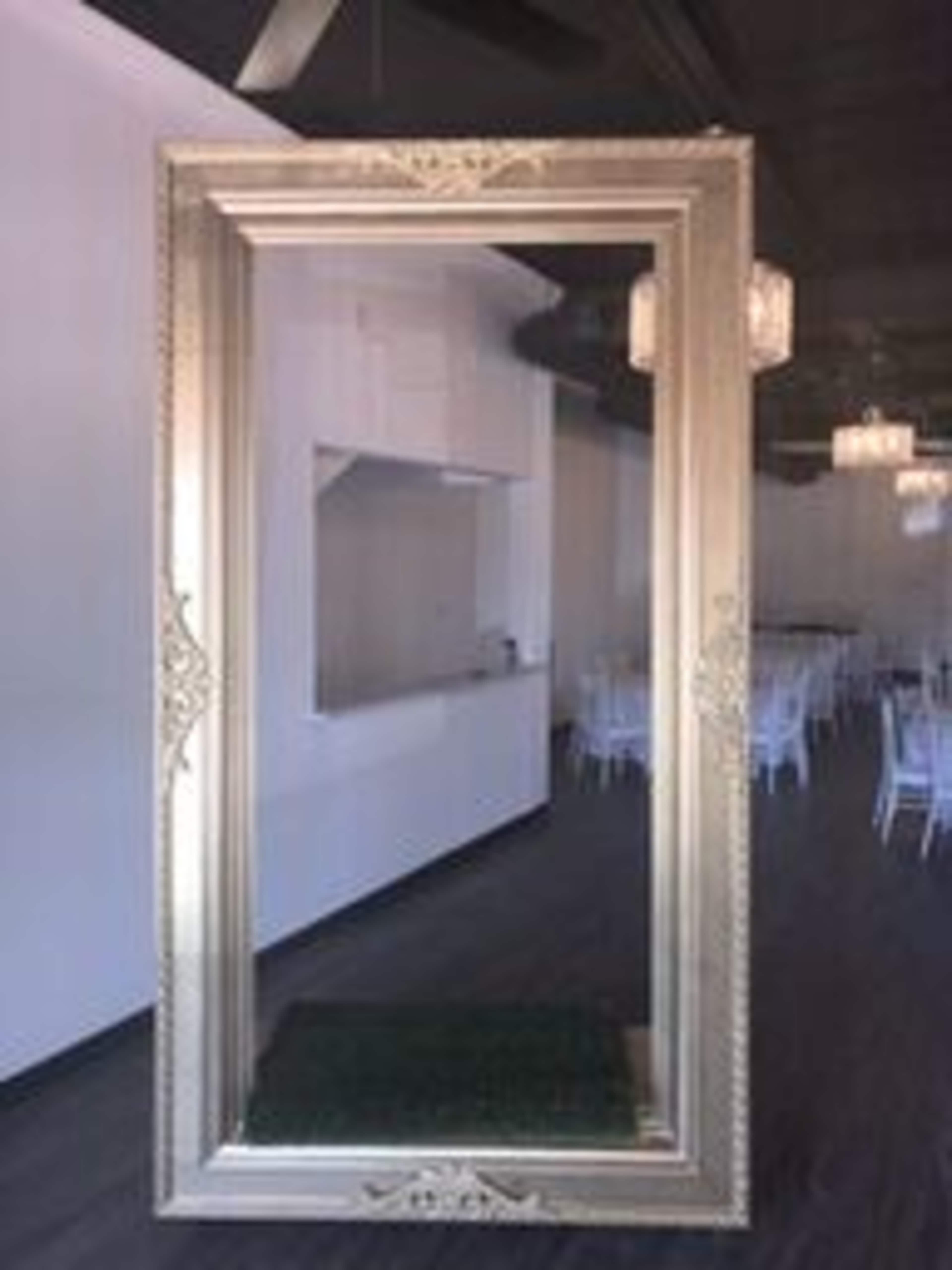 A large, ornate golden picture frame positioned in a modern, brightly lit space with white walls and a visible kitchen area in the background.
