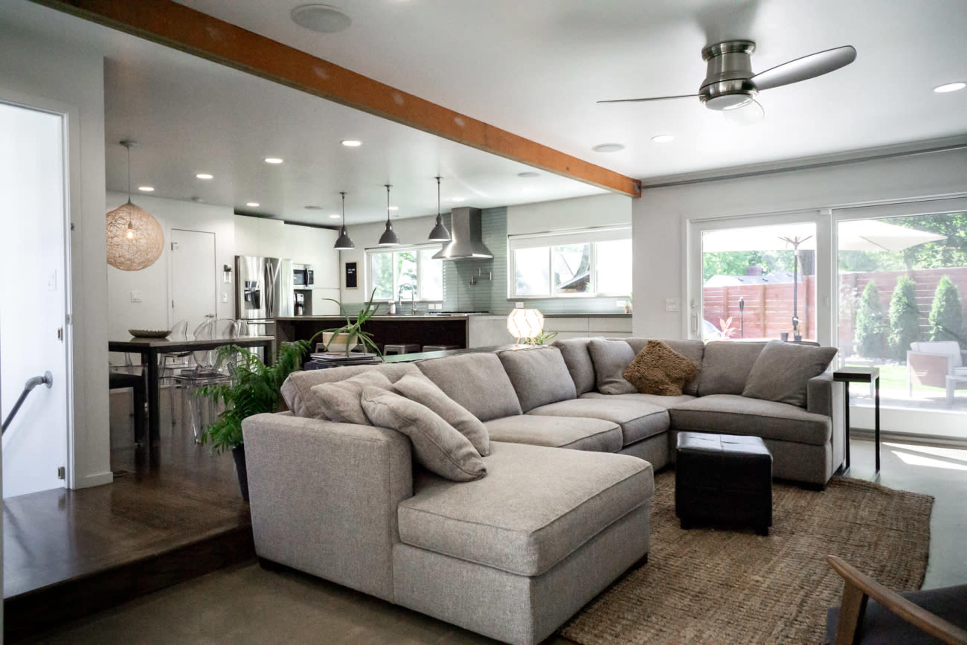 A modern living room with a large sectional sofa, a wooden beam ceiling, and an open view to a kitchen and outdoor patio area.