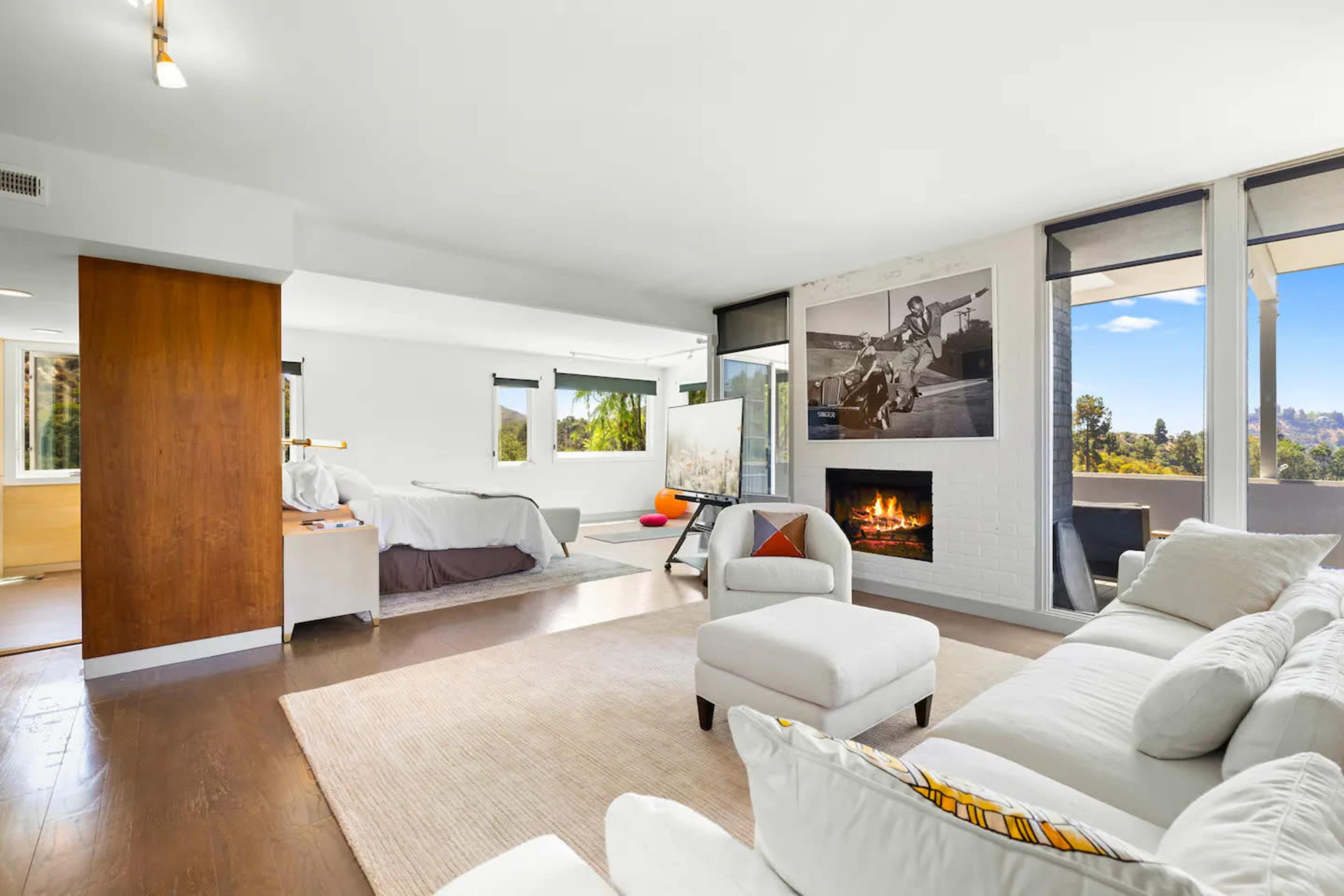 A spacious living room features a white couch, a cozy fireplace, and a large window with a view of outdoor greenery.