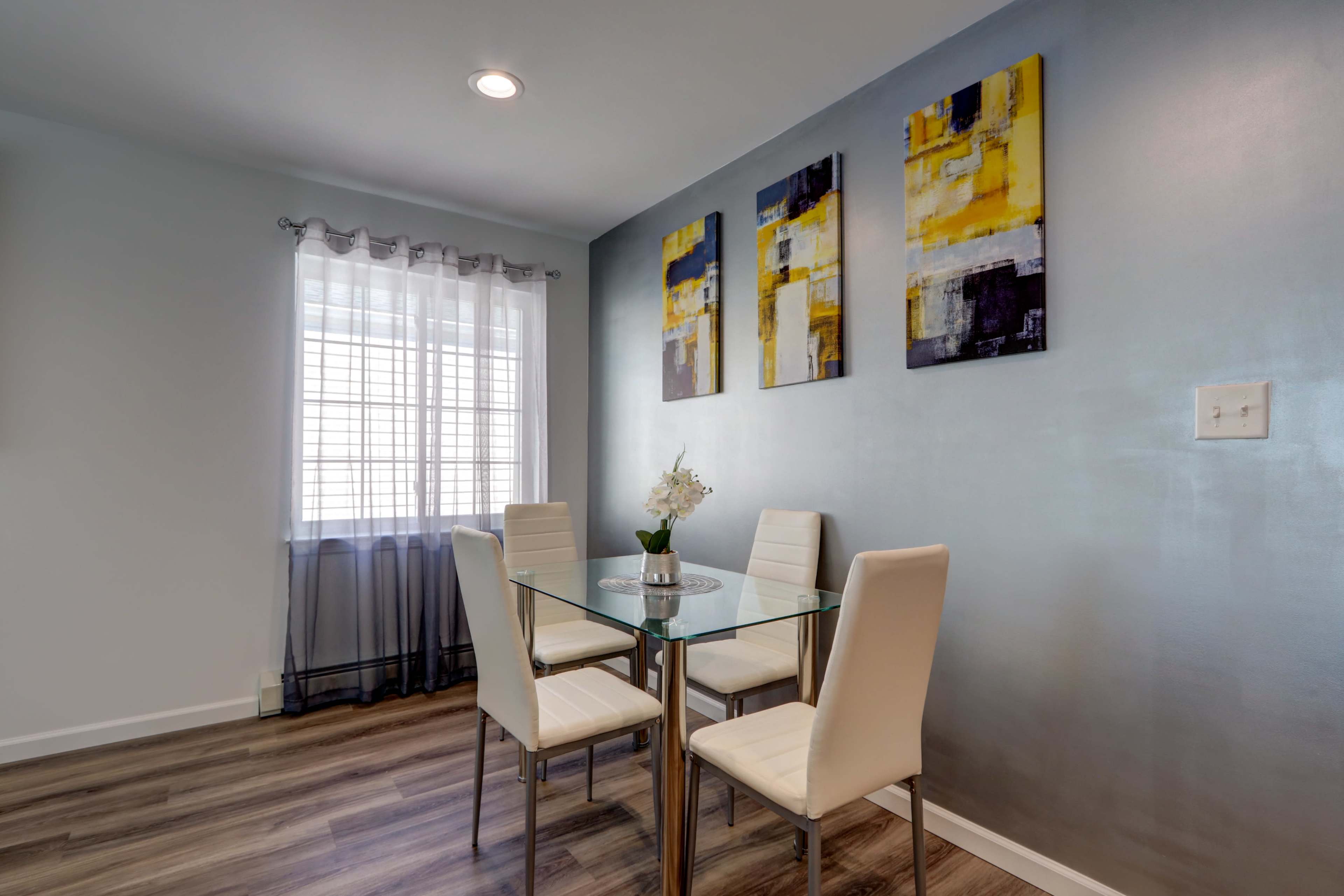 A modern dining area with a glass table and four white chairs, accompanied by abstract artwork on the wall and a window with sheer curtains.