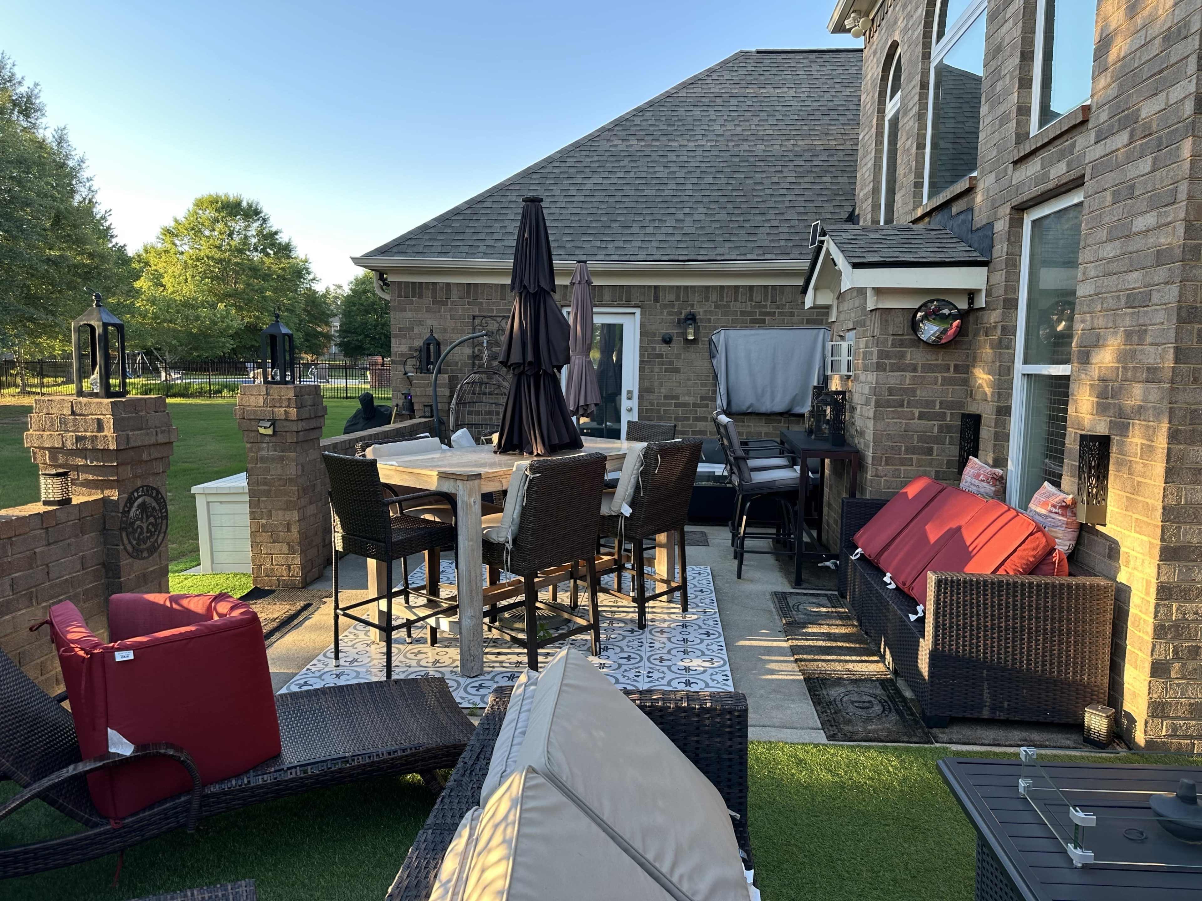 A patio area features outdoor furniture, including a dining table, chairs, and a lounge area, surrounded by greenery and a house wall.
