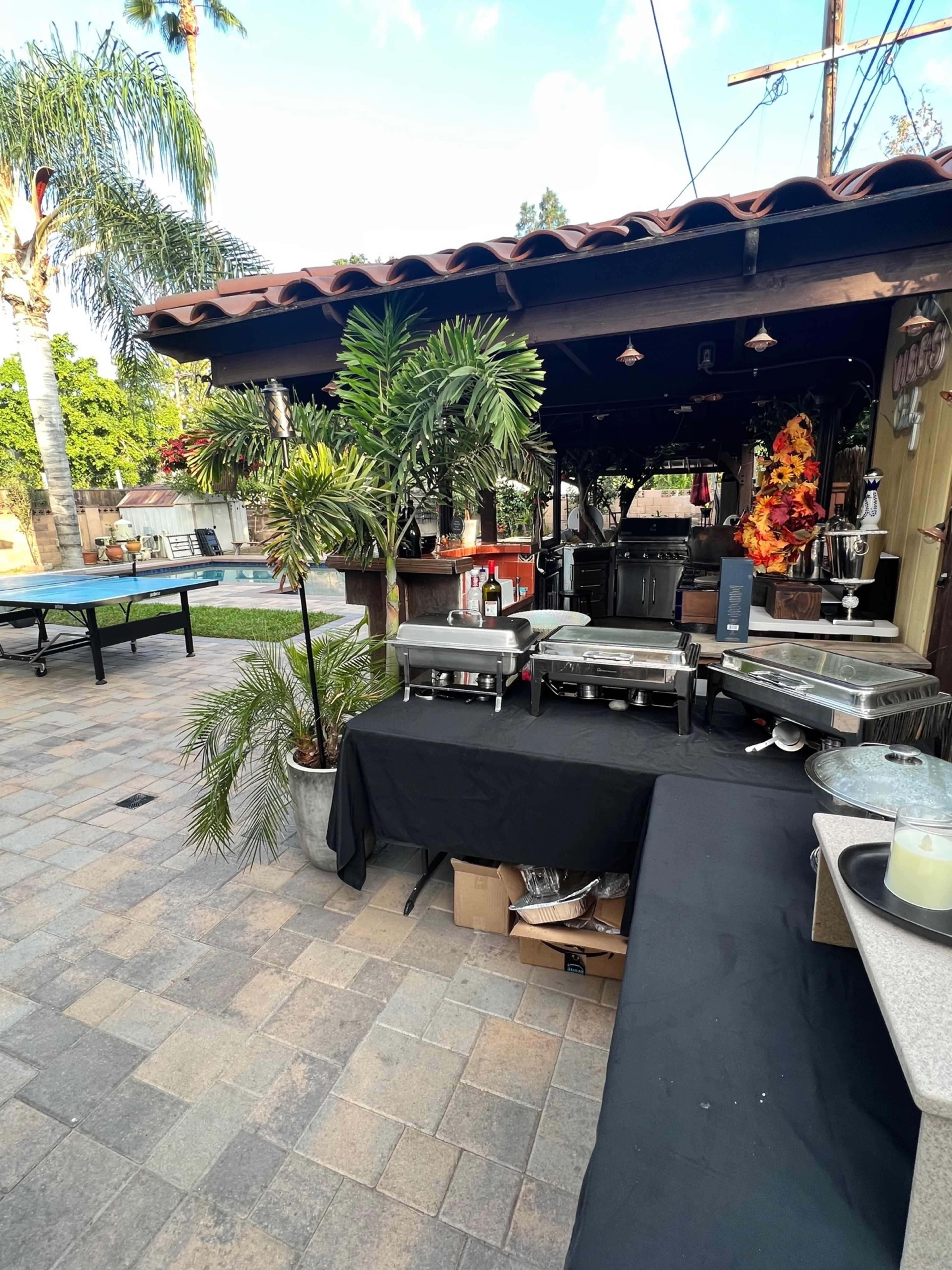 An outdoor area featuring a patio with catering equipment and a pool in the background, surrounded by palm trees.
