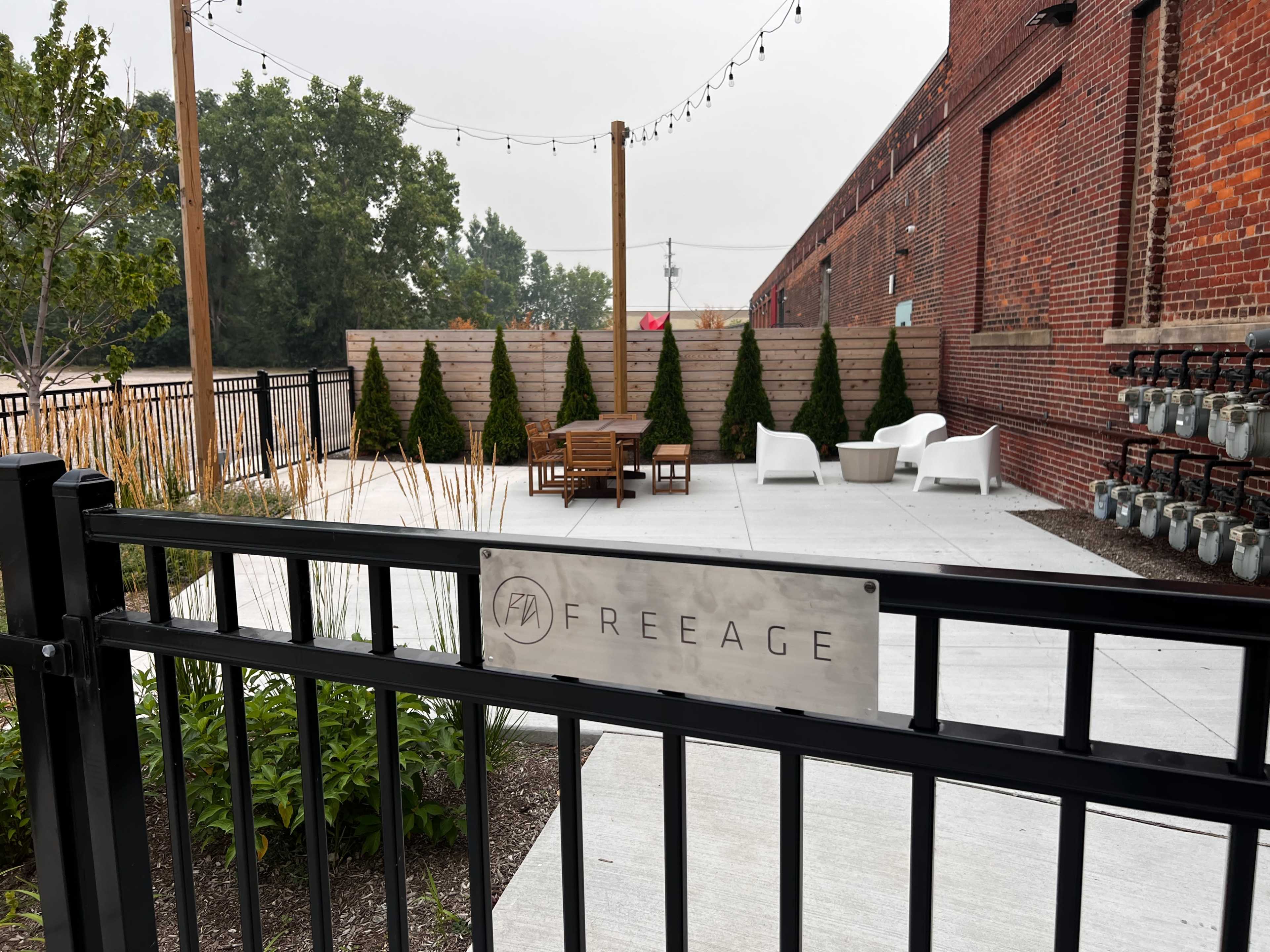 The image shows a fenced outdoor patio area featuring modern seating arrangements and greenery against a backdrop of a brick building.