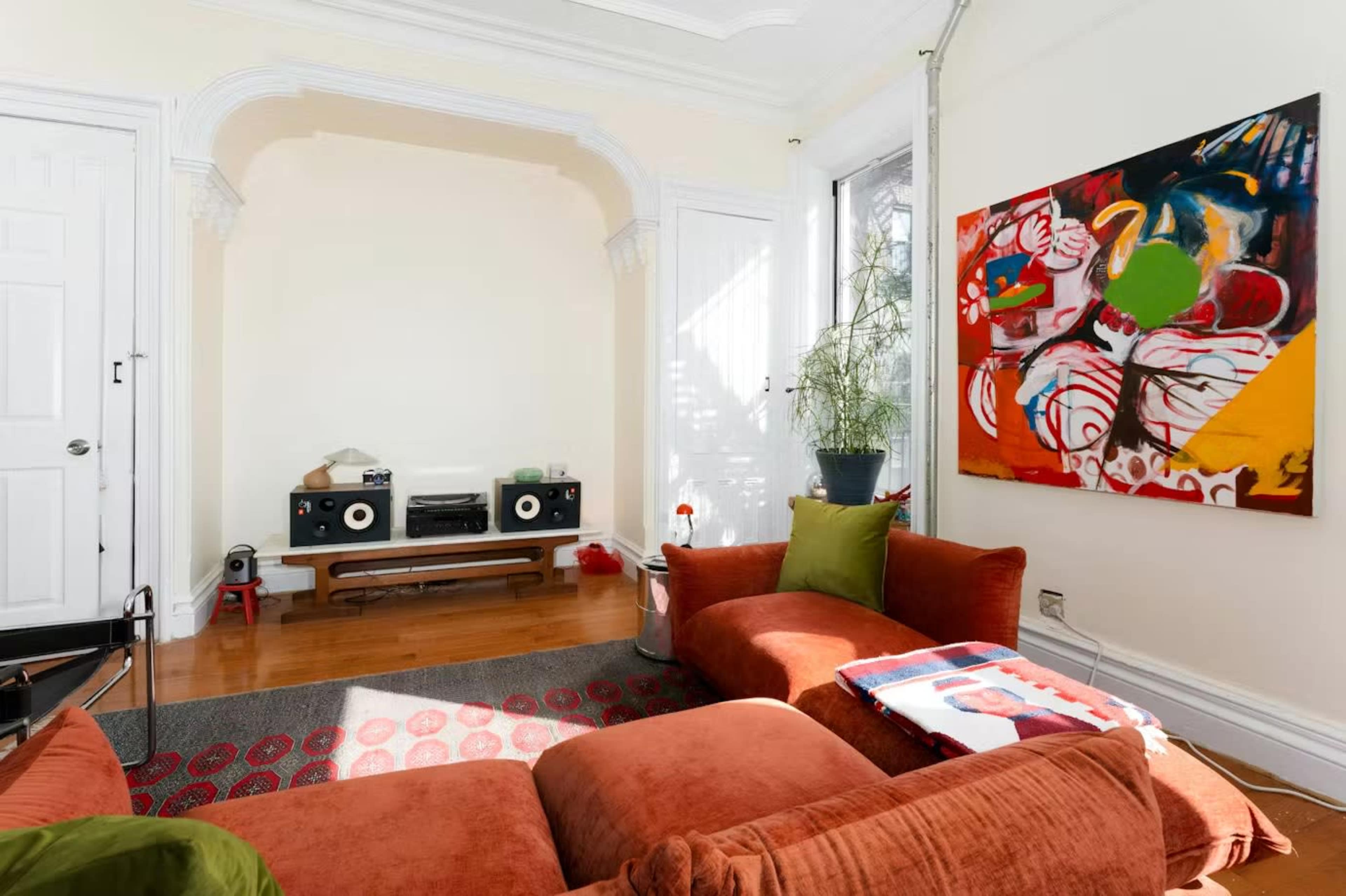 A cozy living room features an orange sectional sofa, a large colorful artwork on the wall, and a vintage audio setup.