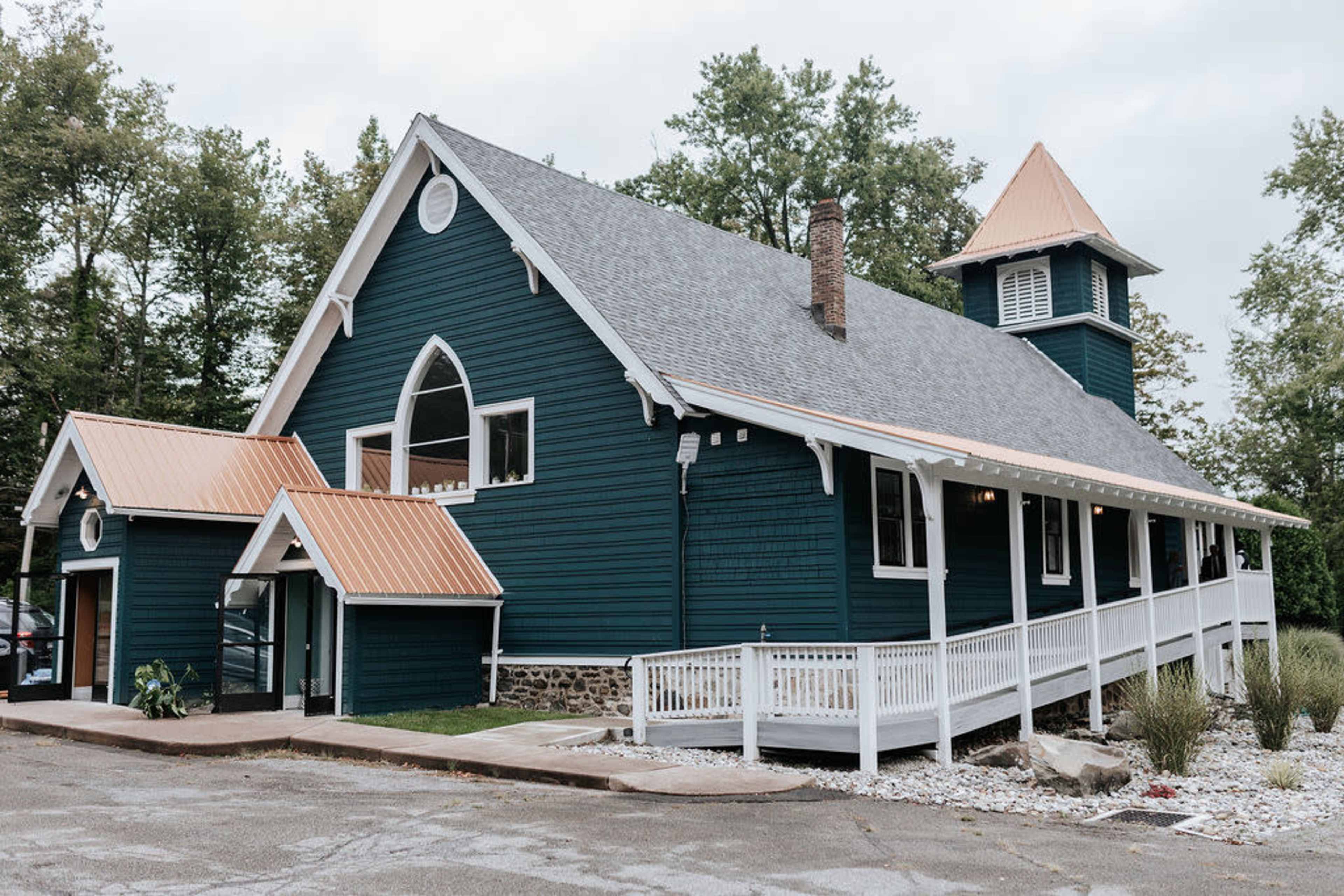 The image shows a large, dark blue wooden building with a peaked roof and a small tower, surrounded by trees and a landscaped area.
