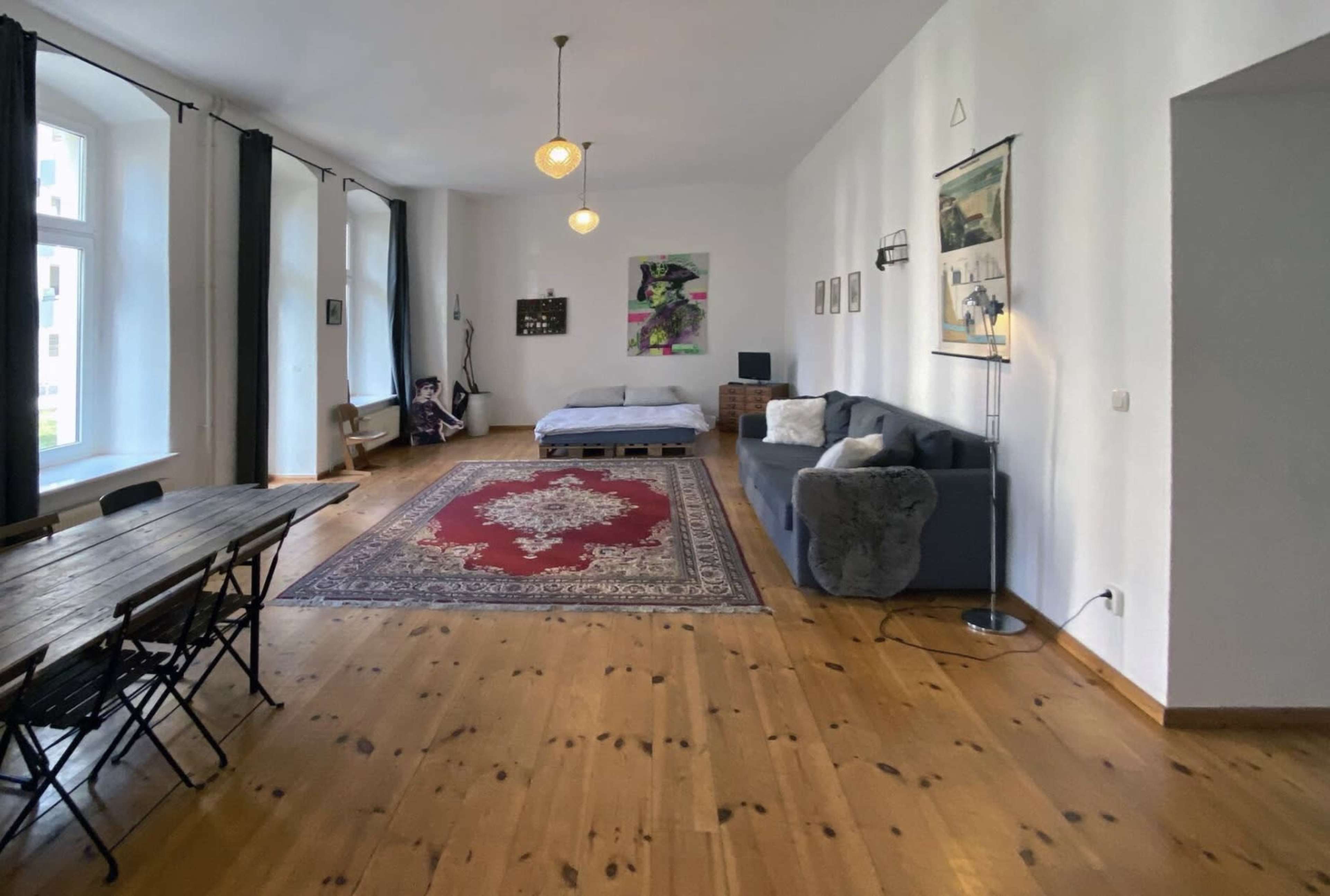 A spacious room with wooden floors, a large area rug, a gray sofa, a bed in the background, and a dining table with chairs.