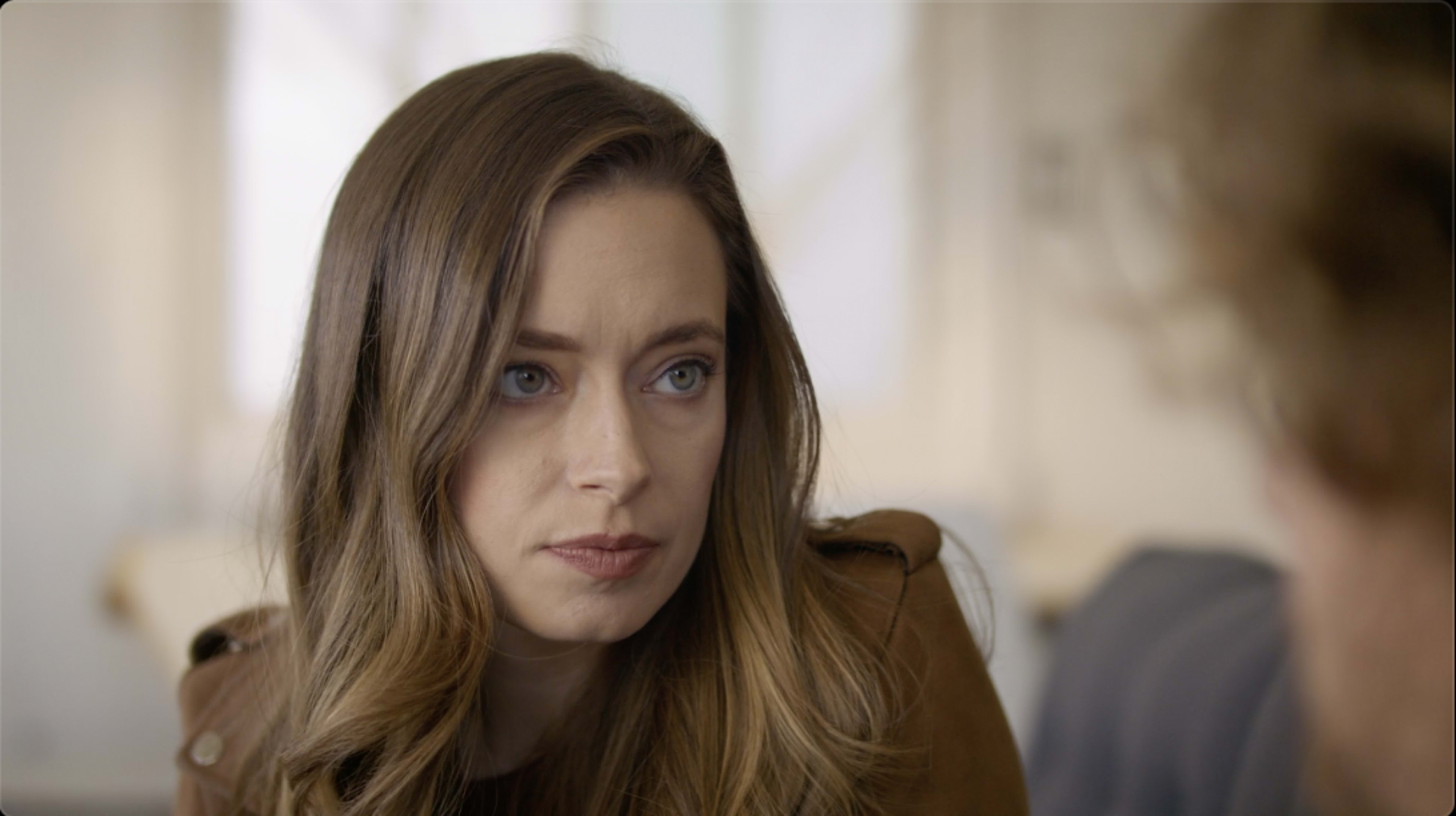A woman with long brown hair and blue eyes listens intently while seated in a bright room.