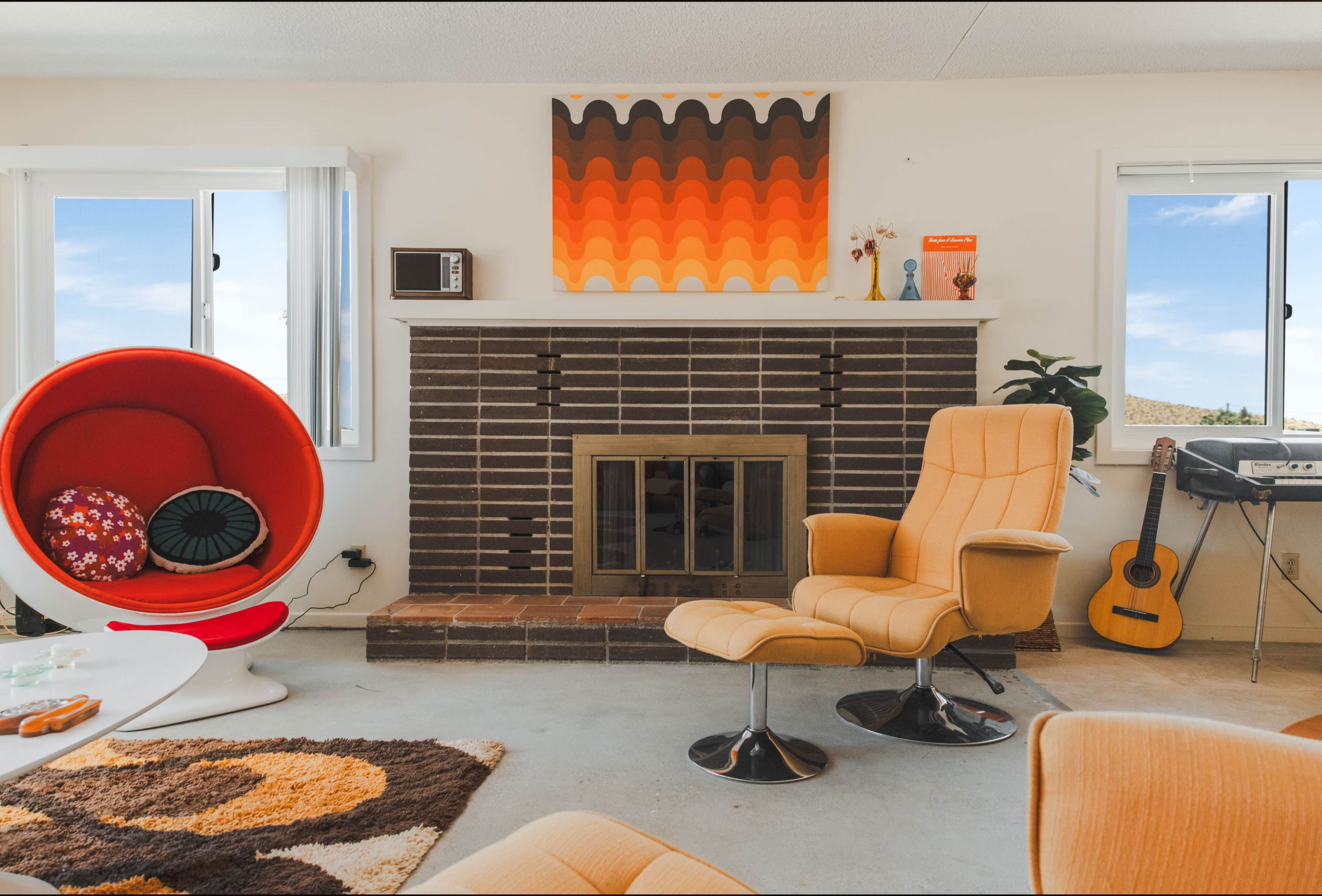 A retro-style living room features a patterned rug, a round red chair, a yellow lounge chair, a brick fireplace, and a wall-mounted television, all accented by colorful wall art.
