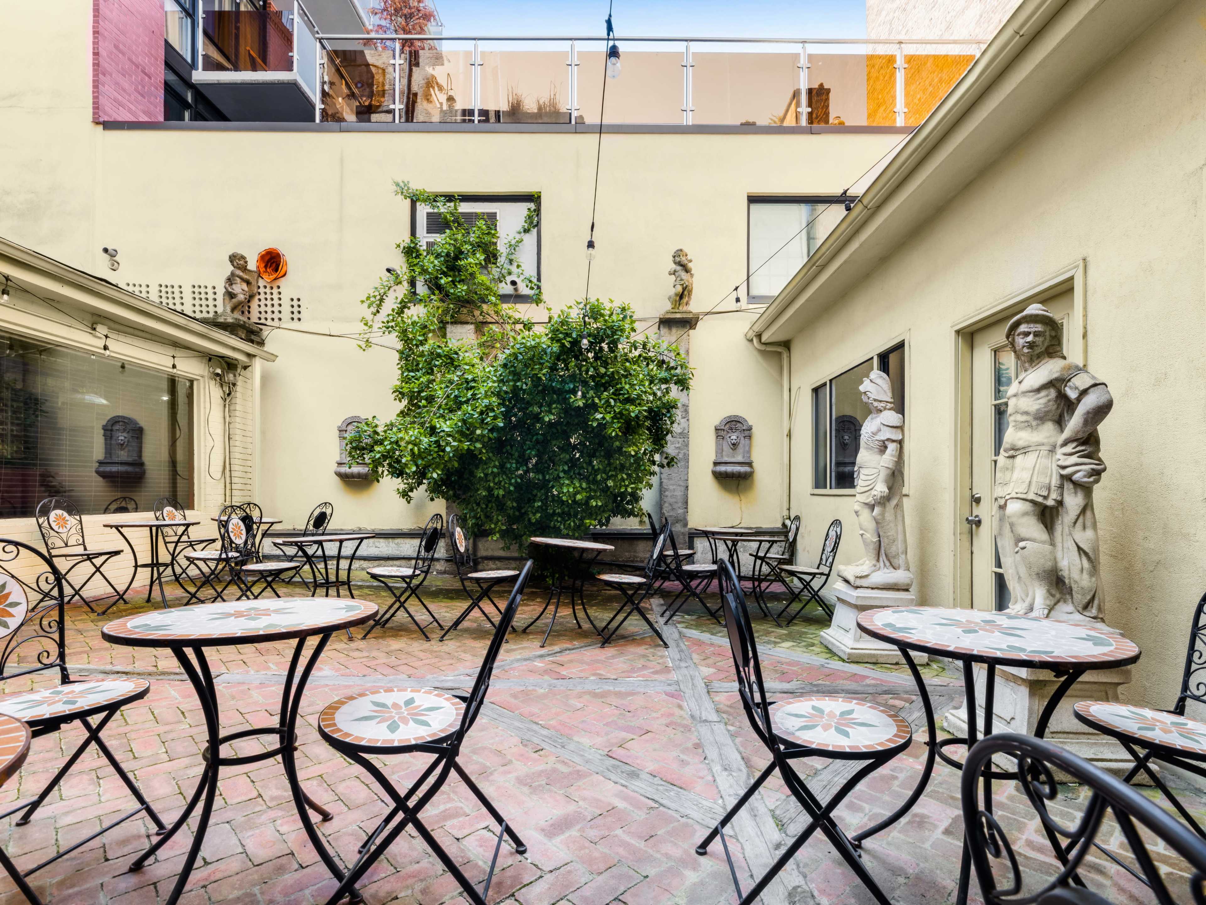 The image shows a courtyard with several metal tables and chairs arranged around a brick floor, with green foliage and statues positioned against the walls.