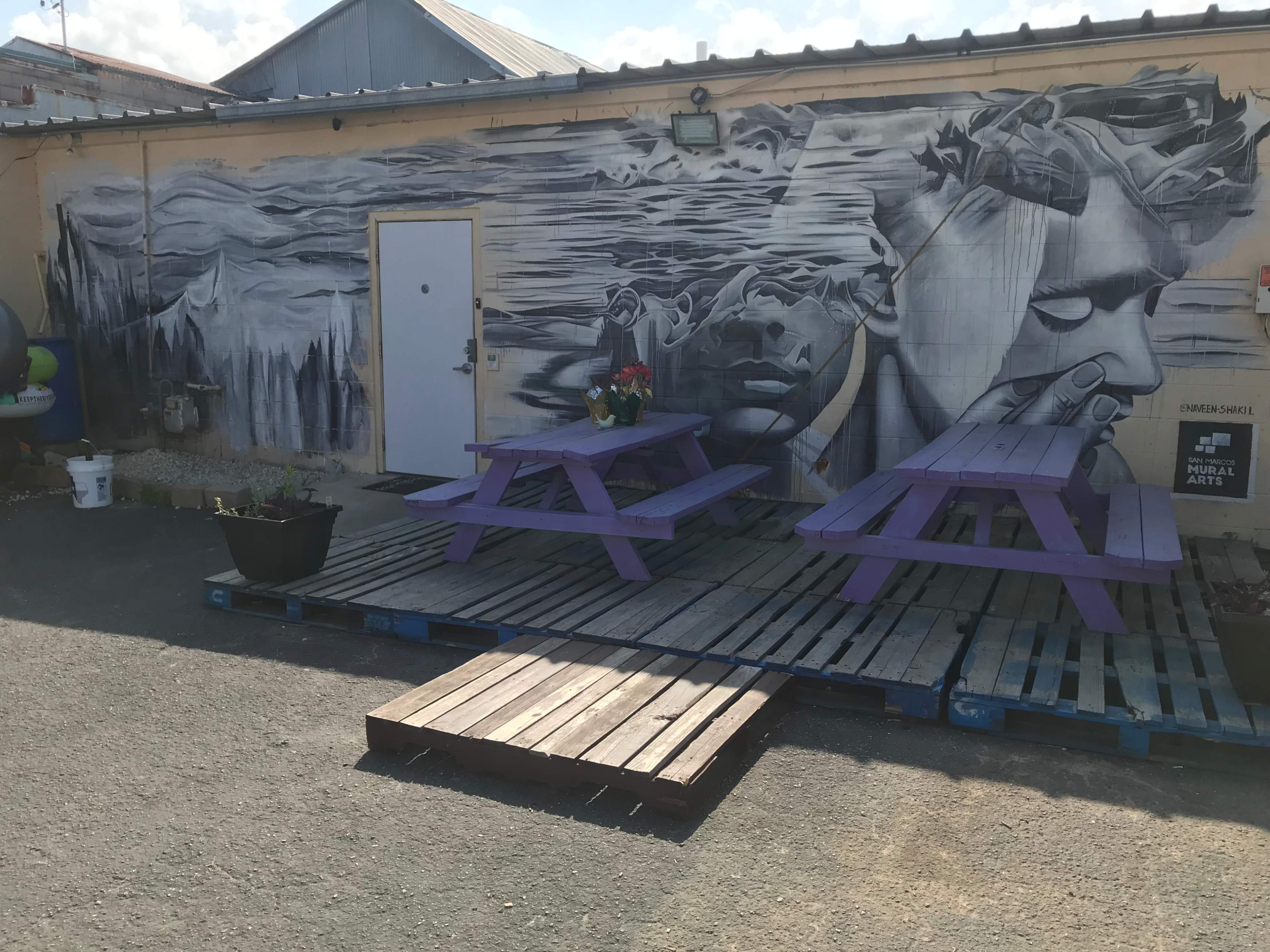 A mural featuring abstract figures on a wall beside two purple picnic tables arranged on wooden pallets in an outdoor area.