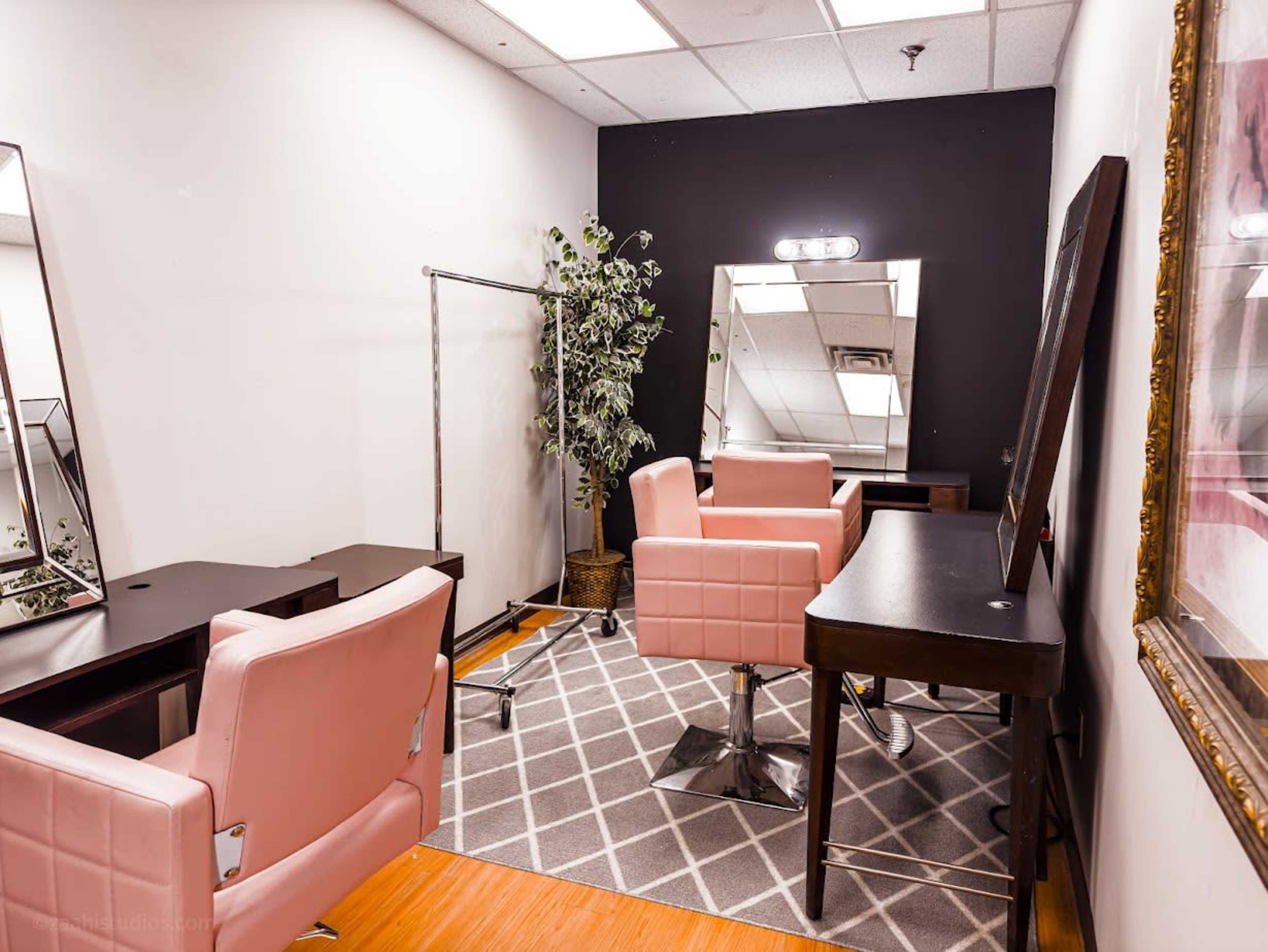 A modern salon interior with two pink styling chairs, a mirror, and a clothing rack against a black wall.
