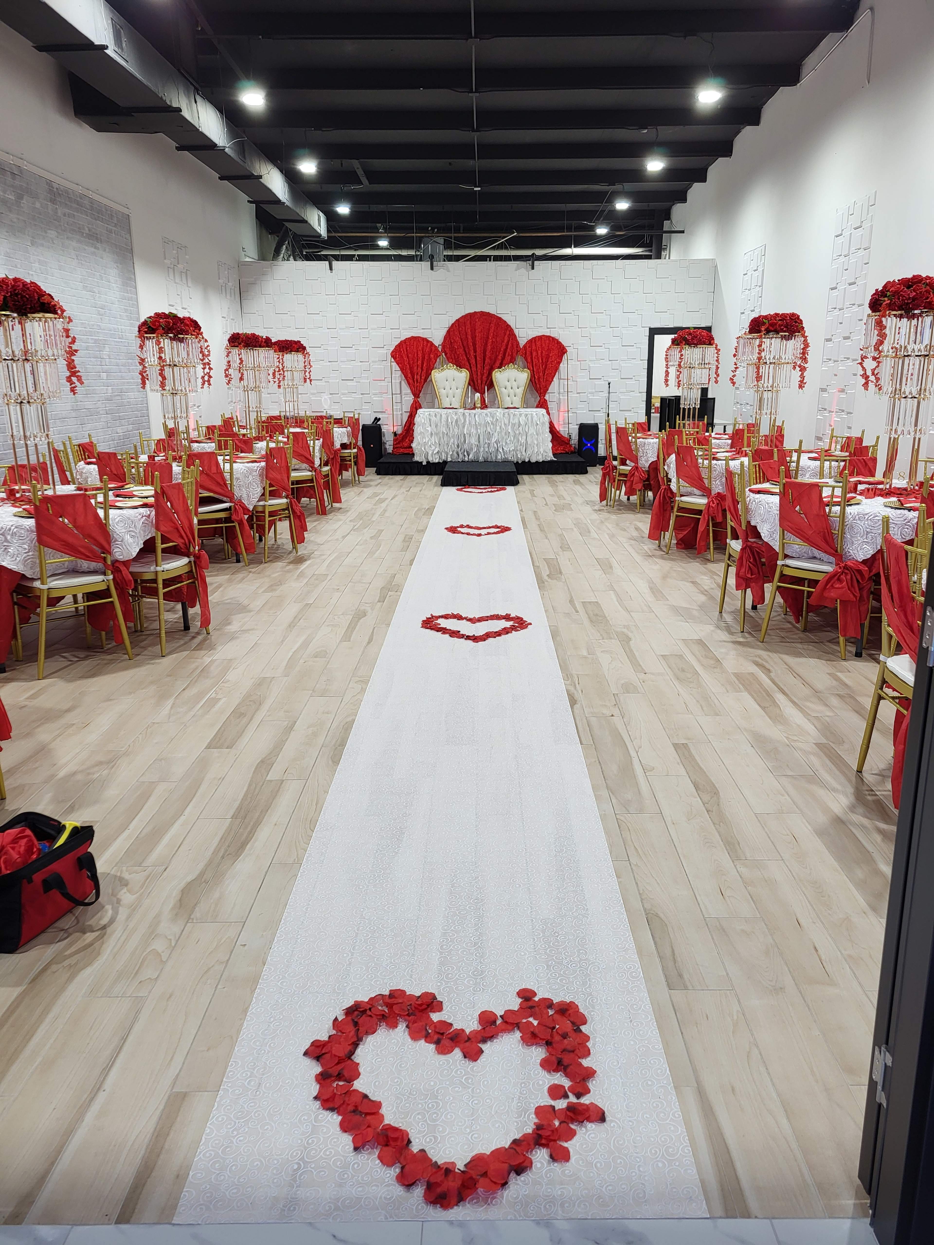 The image depicts a banquet hall decorated for an event with red and white themes, featuring tables adorned with red tablecloths and centerpieces, and a white aisle runner sprinkled with red rose petals arranged in heart shapes.
