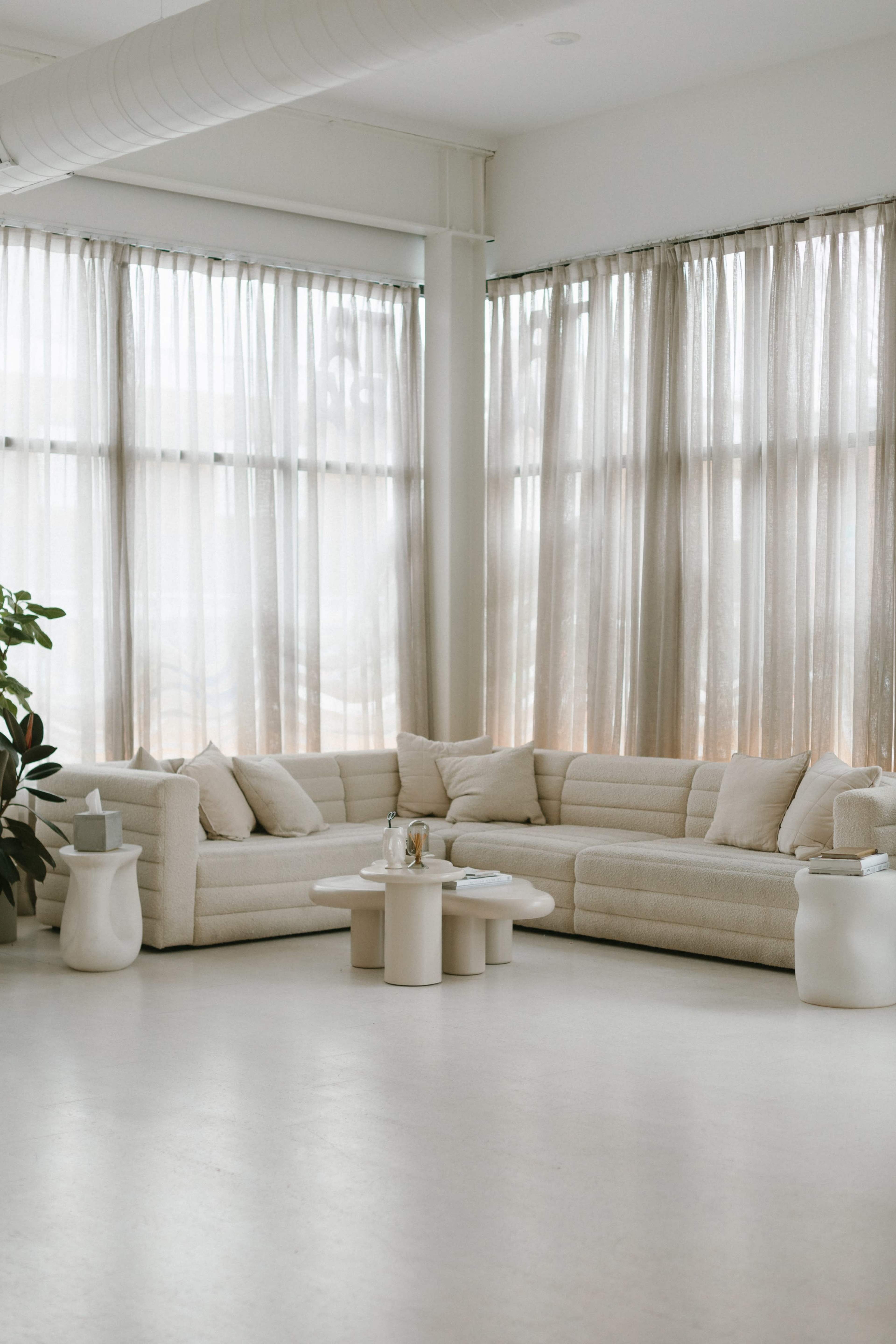 A light-filled room features a spacious beige sectional sofa, a circular coffee table, and sheer curtains.