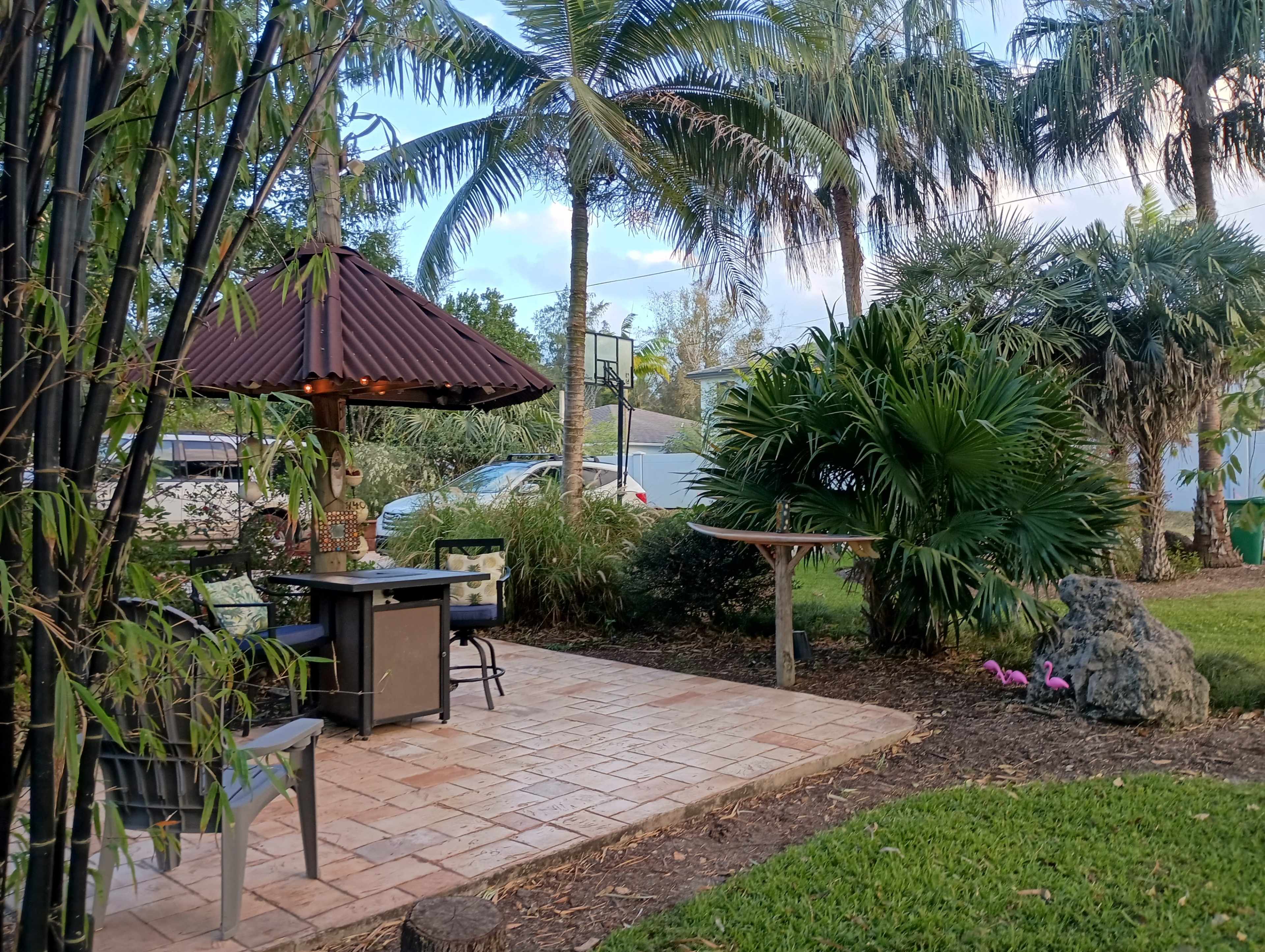 The image shows a patio area with a gazebo, outdoor seating, and lush tropical landscaping featuring palm trees and a rock formation.
