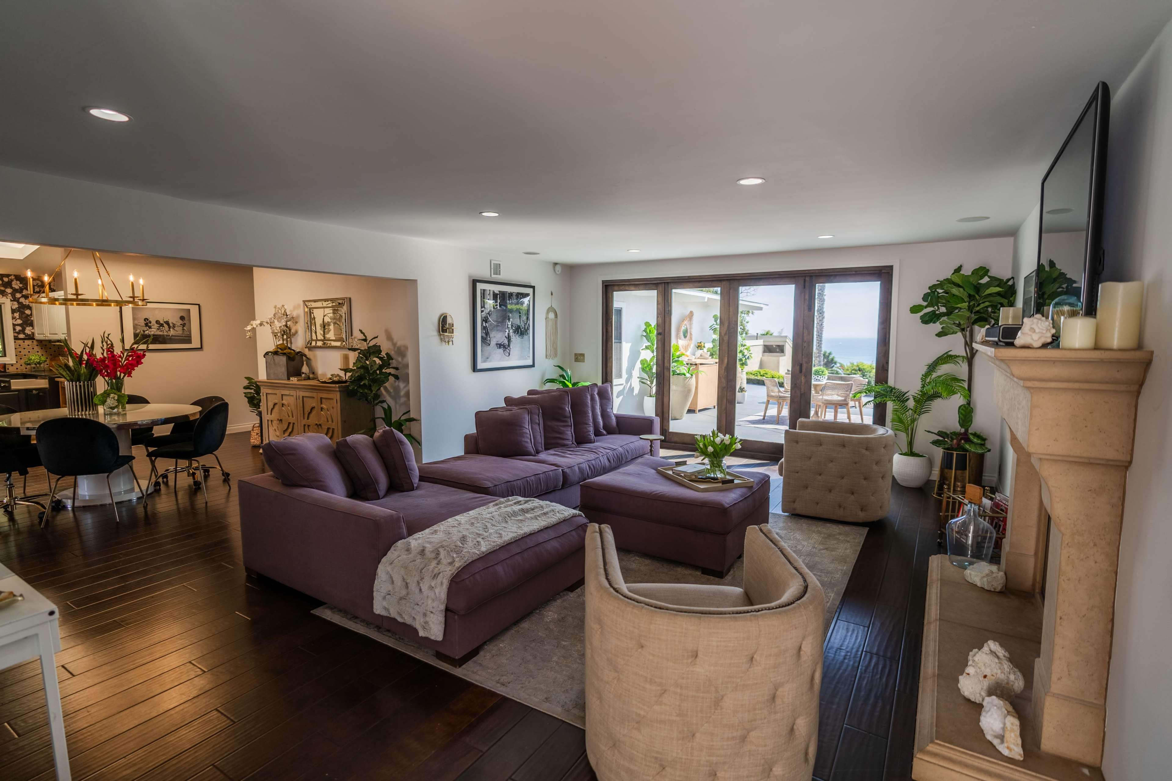 The scene shows a modern living room with purple sofas, a coffee table, and large windows that open to a patio with a view.