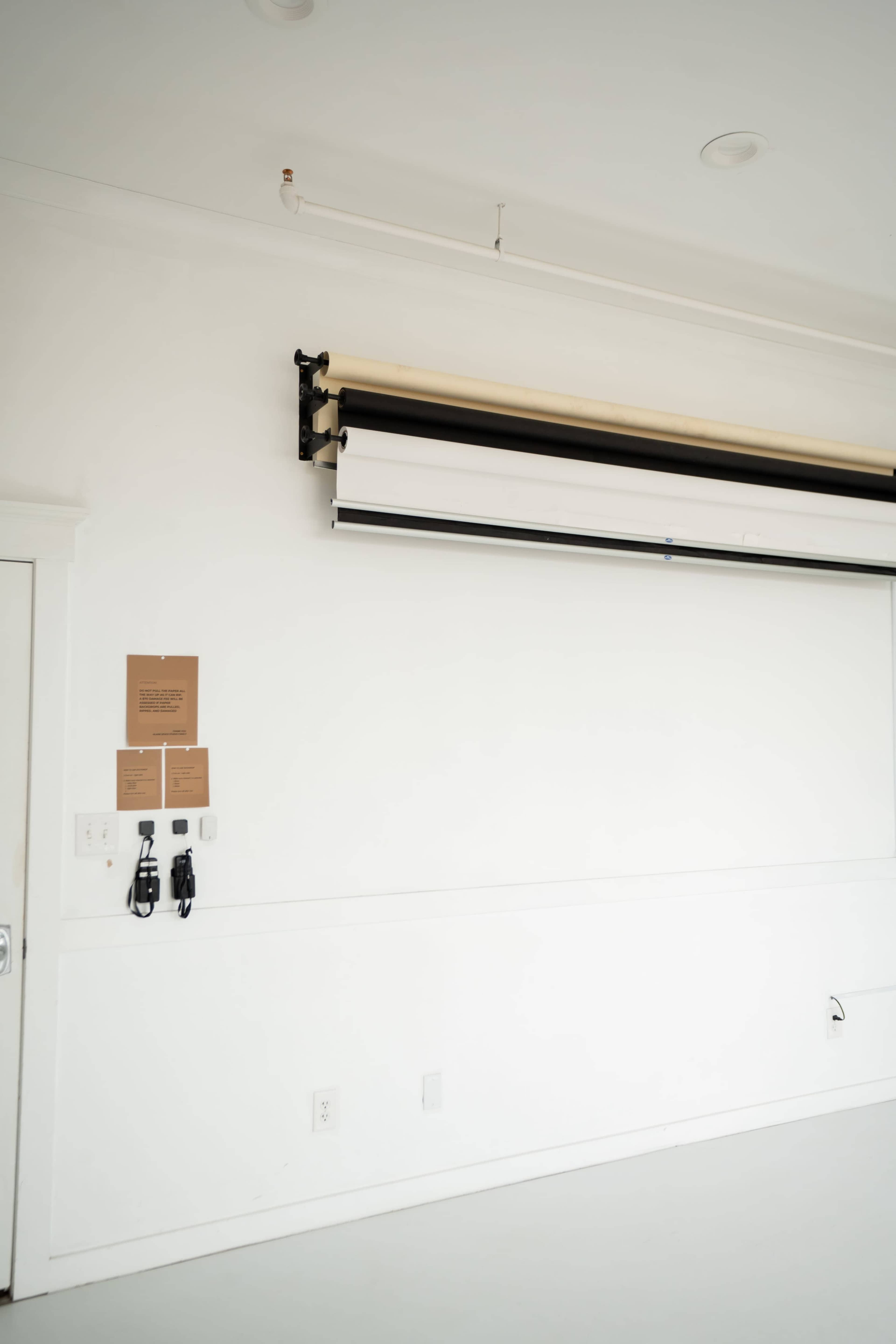 The image shows a minimalistic room with a white wall, a roll-down backdrop system mounted above, and a door on the left.