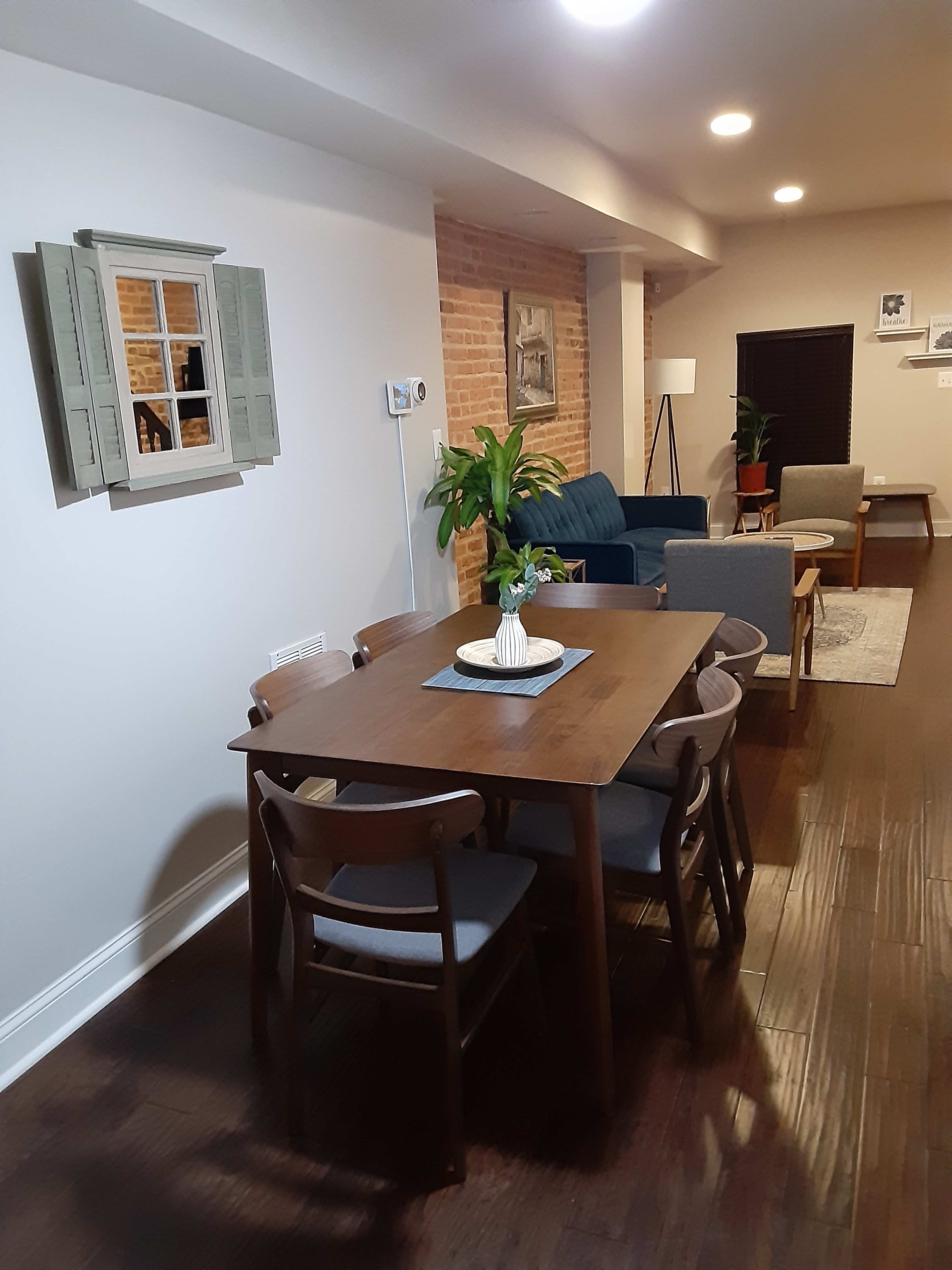 A wooden dining table with six chairs is positioned in a well-lit room featuring a brick accent wall and a cozy seating area.
