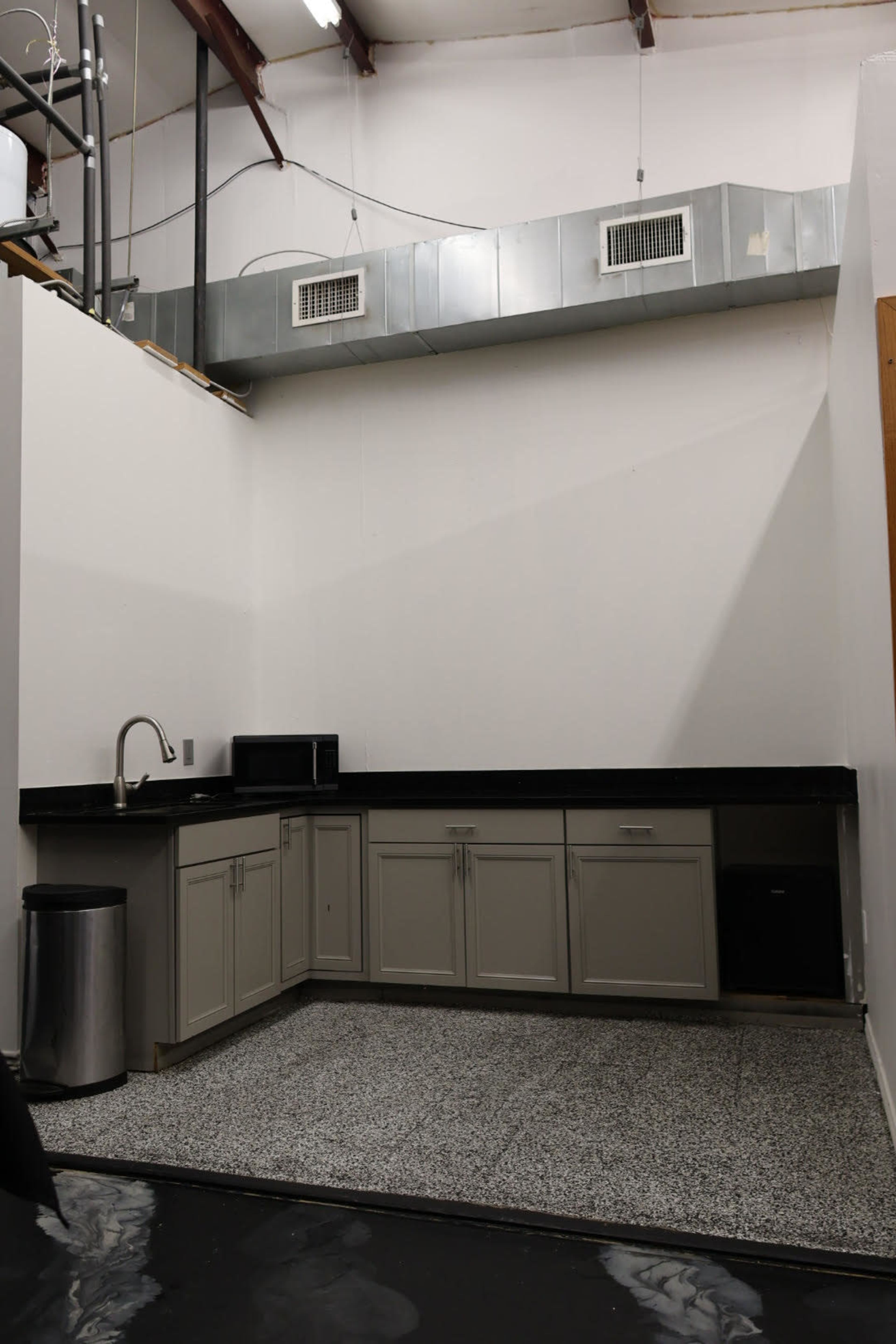 The image shows a small kitchen area featuring a black countertop, a sink, a microwave, and cabinets, with a gray floor and venting above.