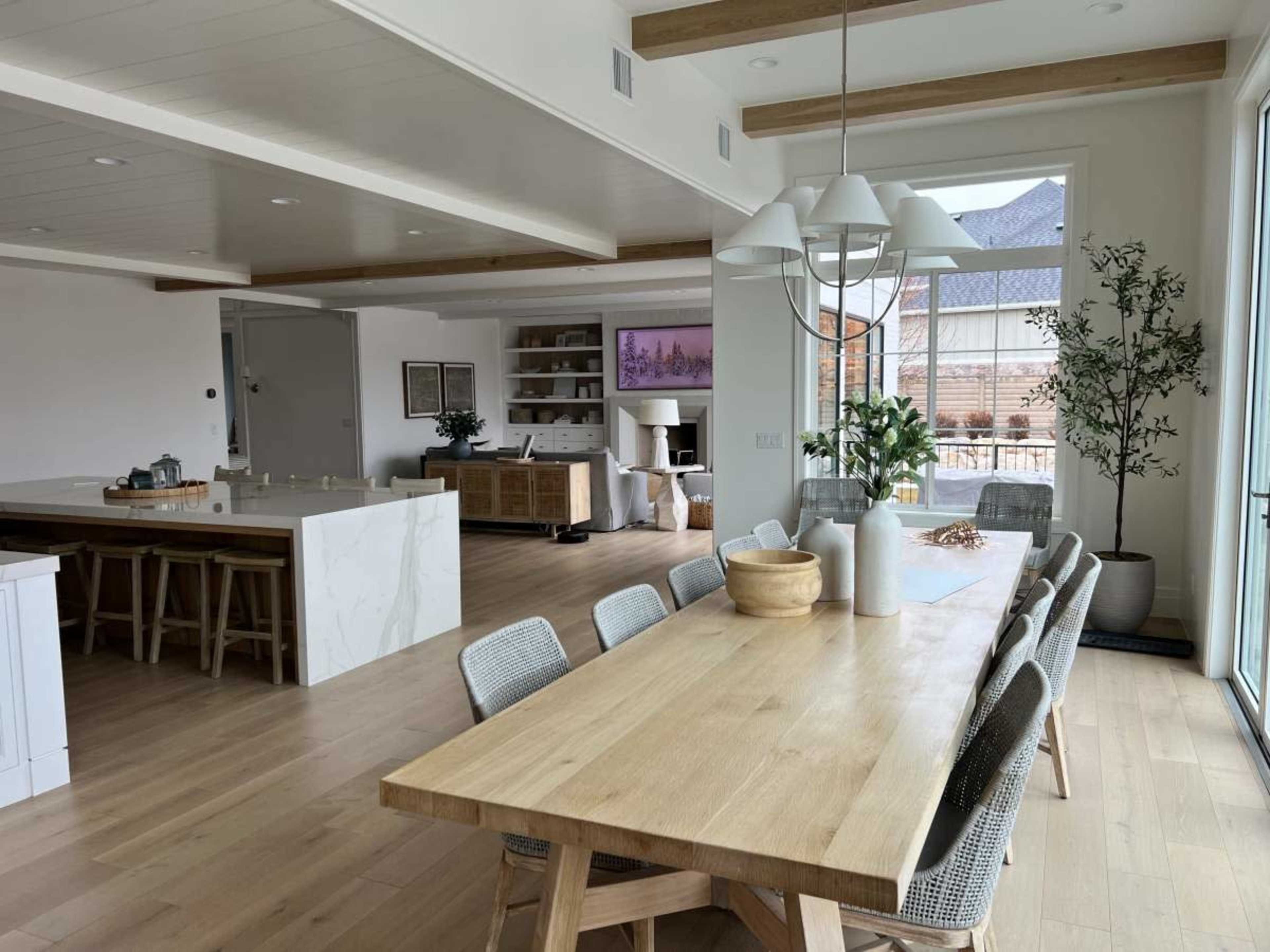 The image shows a modern open-concept kitchen and dining area with a large wooden dining table, light-colored cabinetry, and a spacious living room visible in the background.